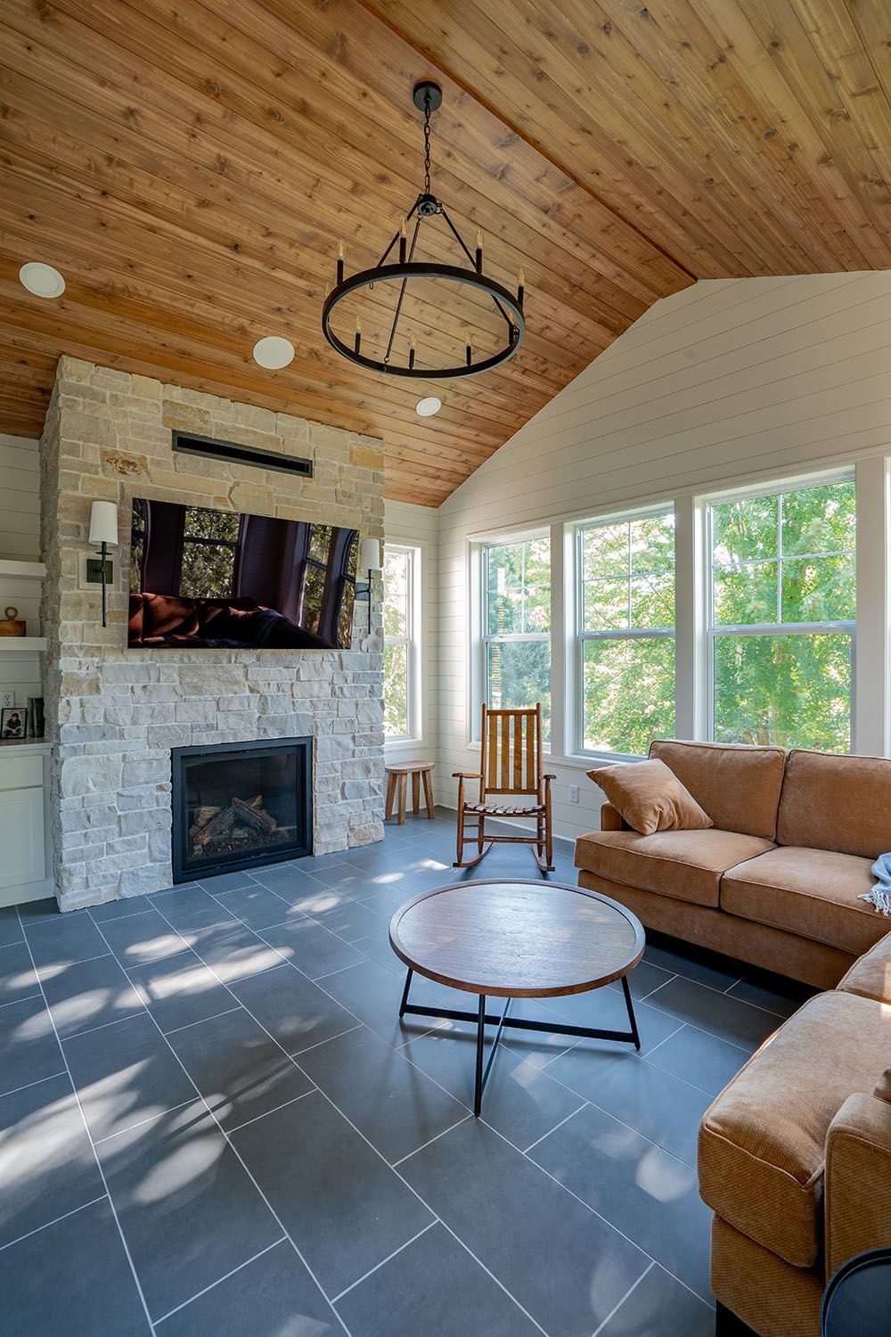 Cozy sunroom with stone fireplace, tan sectional, round coffee table, and large windows under a wood ceiling