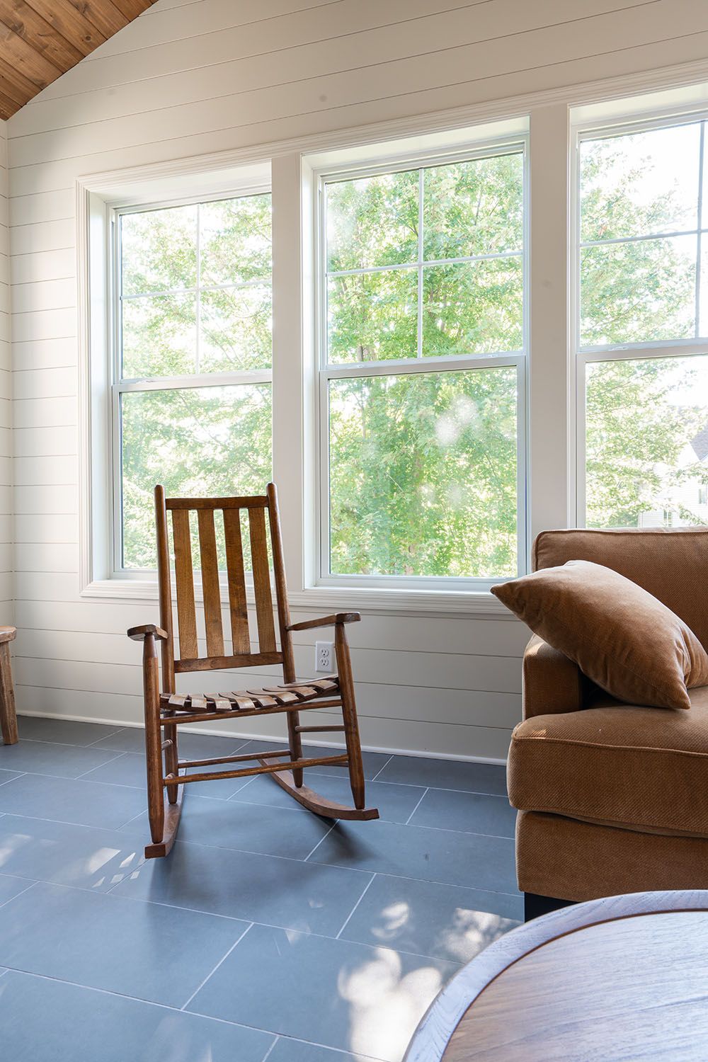 Sunlit living room with large windows, a wooden rocking chair, and a brown sofa with pillows