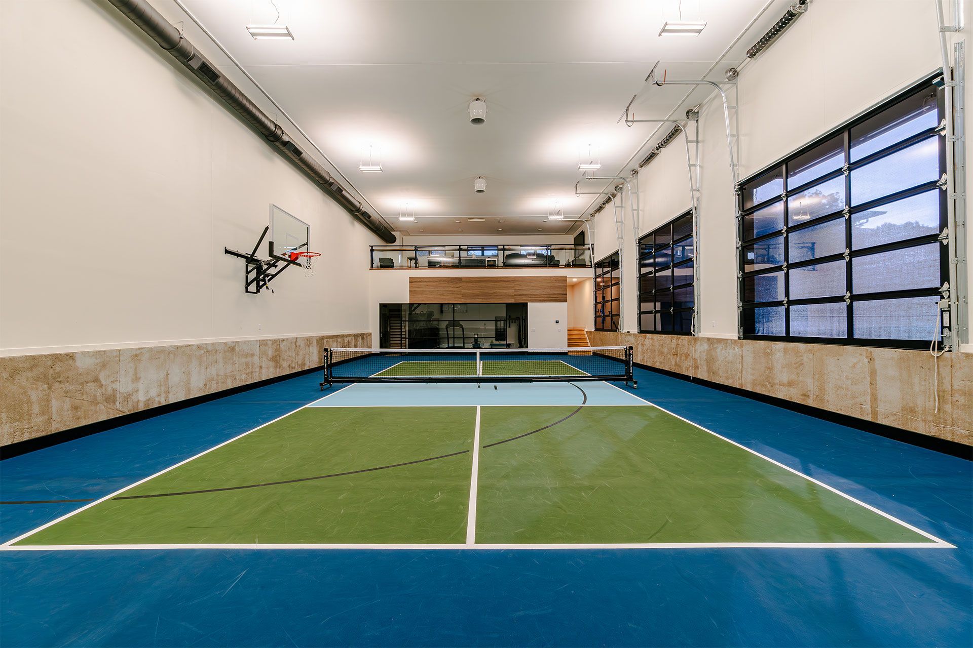 Indoor pickleball court with blue and green flooring, net, basketball hoop, and windows.