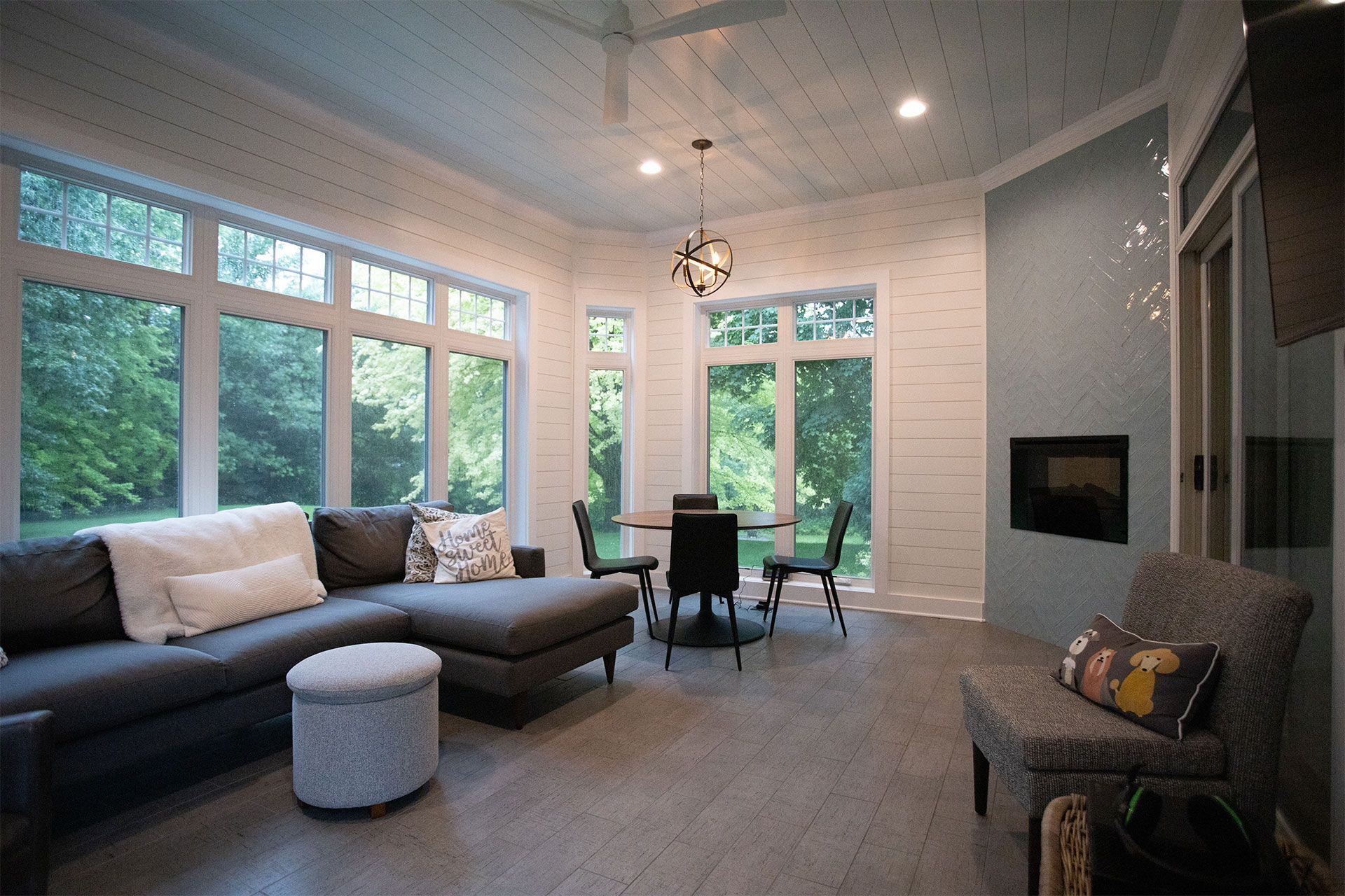 Sunroom with large windows overlooking trees, sectional sofa, round table with chairs, and fireplace.