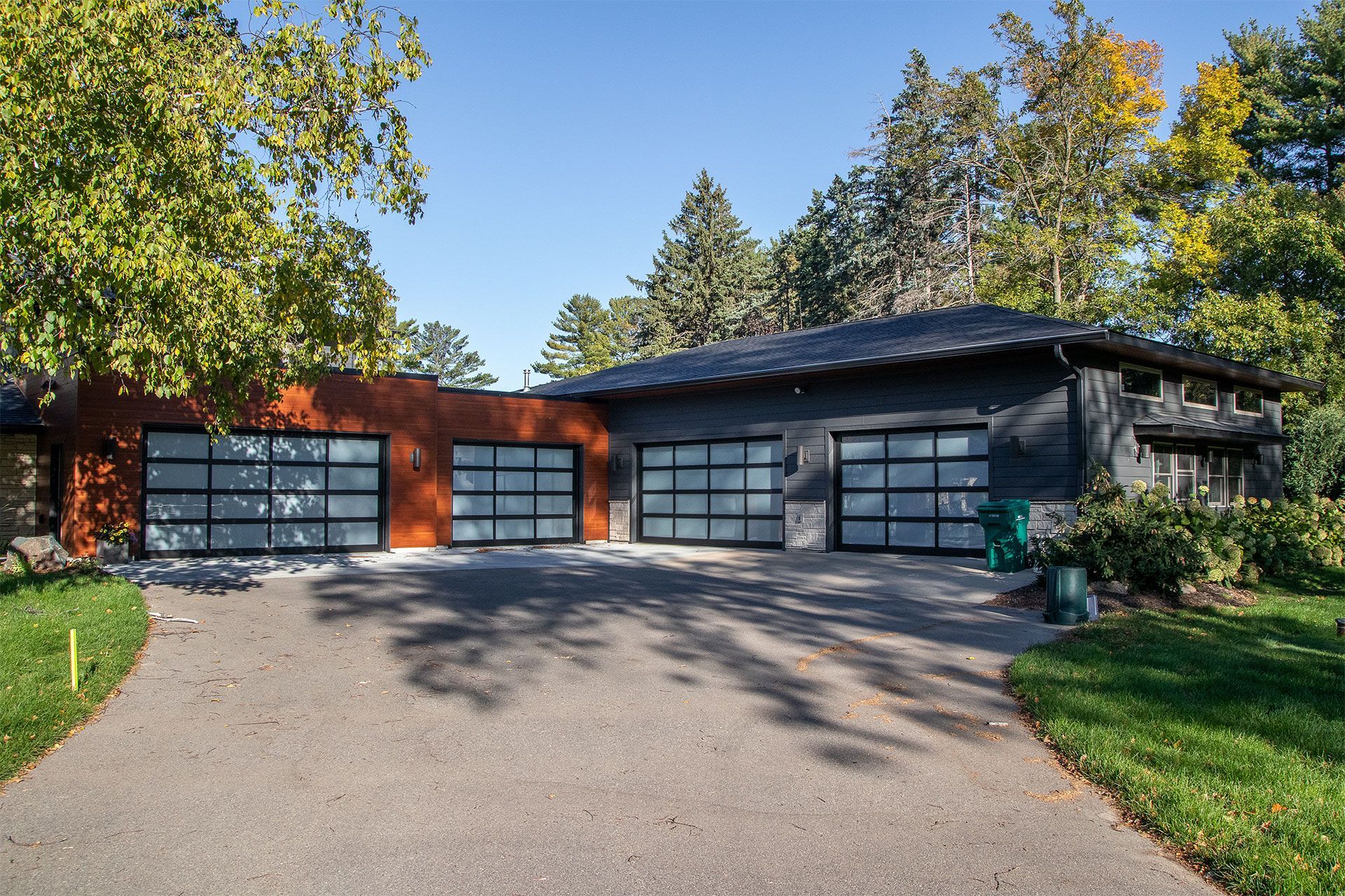Modern home with three-car garage, glass doors, and dark gray siding. Brown and green accents, driveway, sunny day.