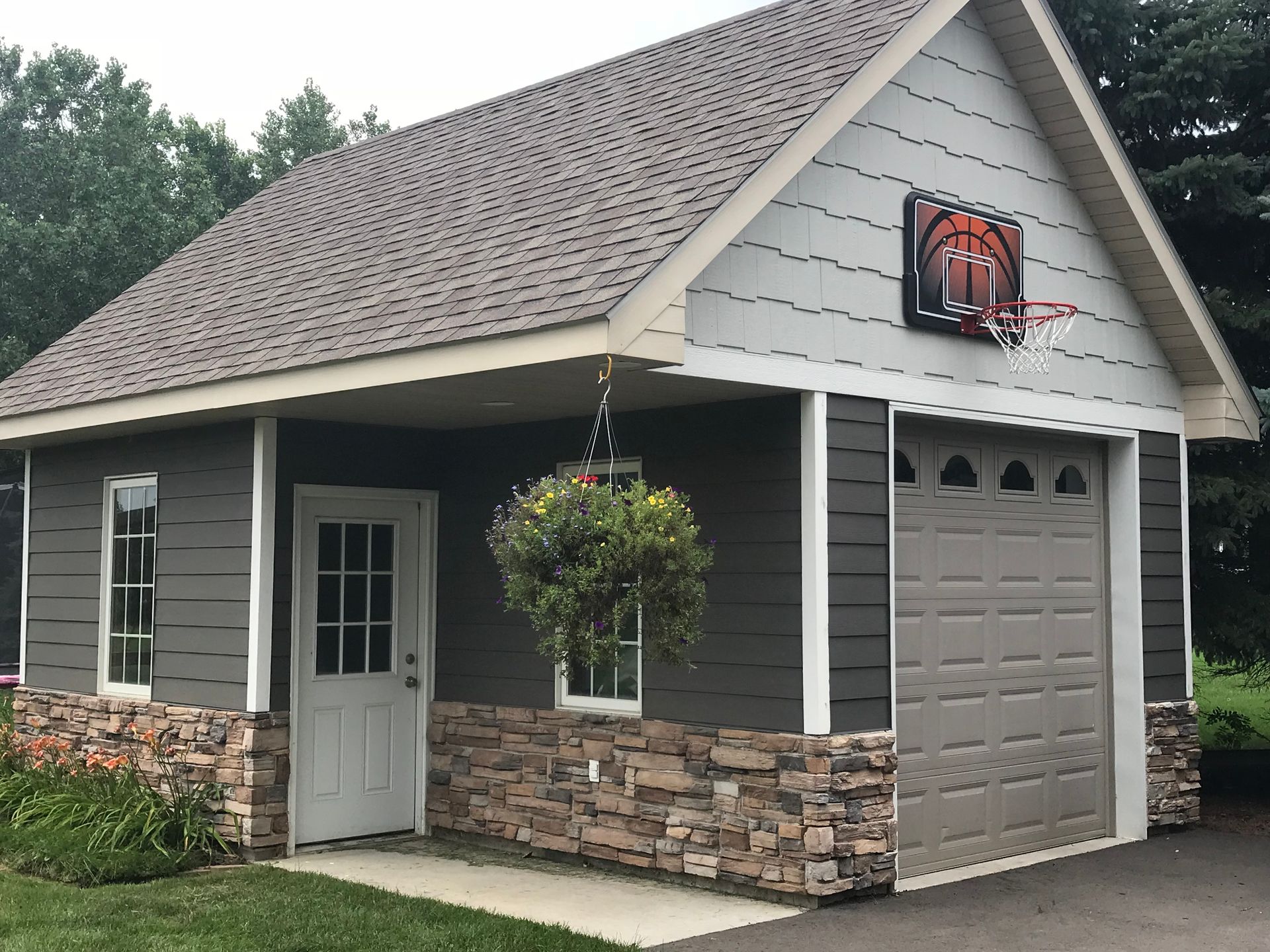 Garage with gray siding, stone base, brown roof, basketball hoop, hanging basket.