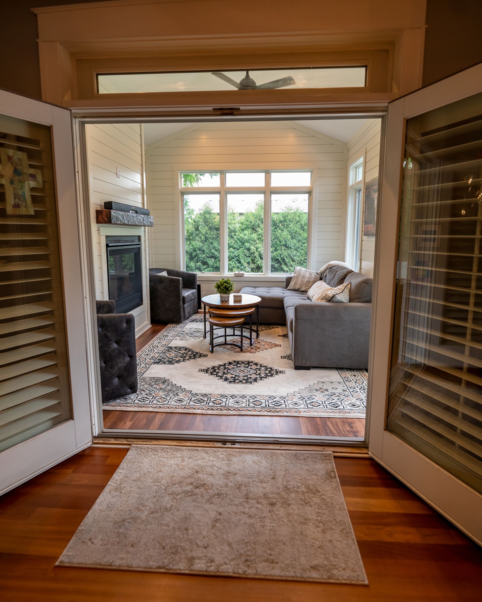 Open doors lead to a sunroom with gray couches, rug, coffee table, and large windows with greenery.