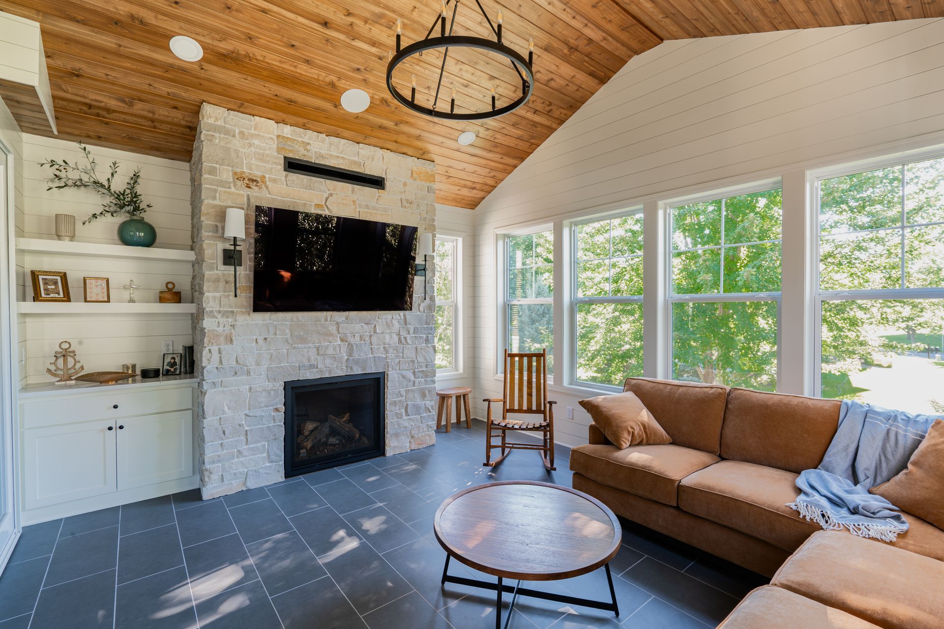 Sunroom with a stone fireplace, brown leather sectional, wooden ceiling, and large windows.