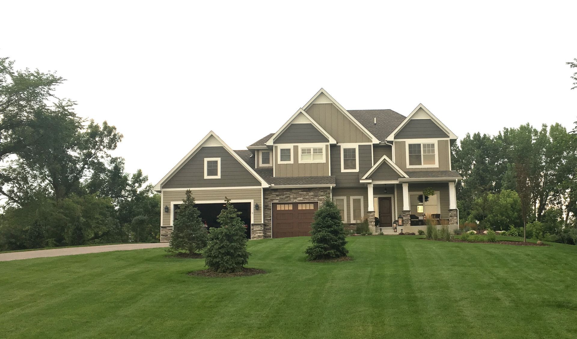 Large two-story house with green lawn, trees, and gray and brown siding under a cloudy sky.