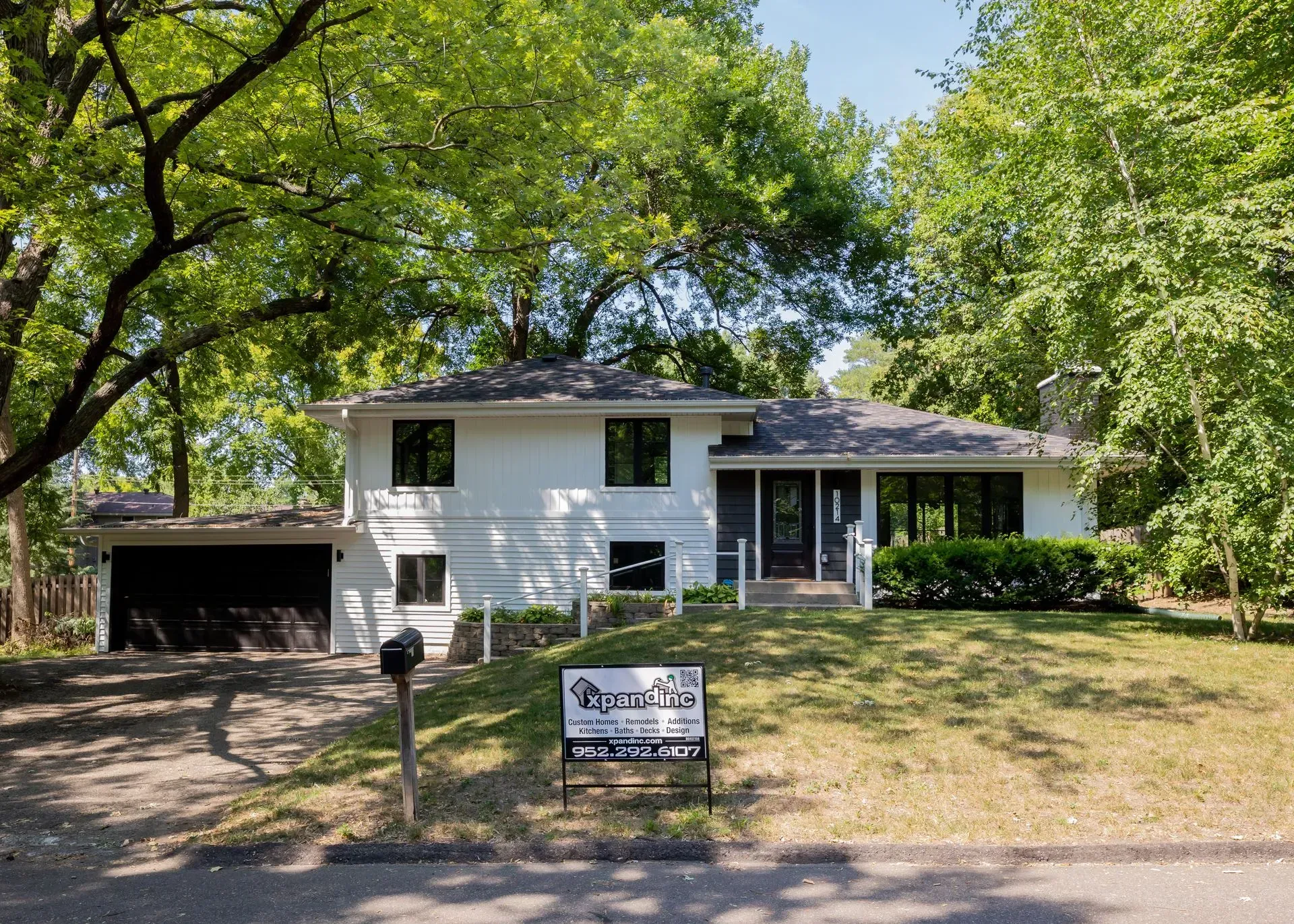 White house with black trim, surrounded by trees, lawn sign in front.
