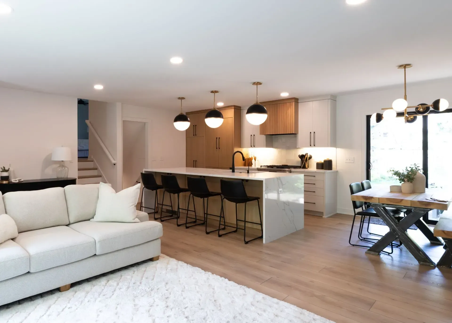 Open-concept kitchen and dining area with island seating, black chairs, and light wood cabinetry, with living room visible.