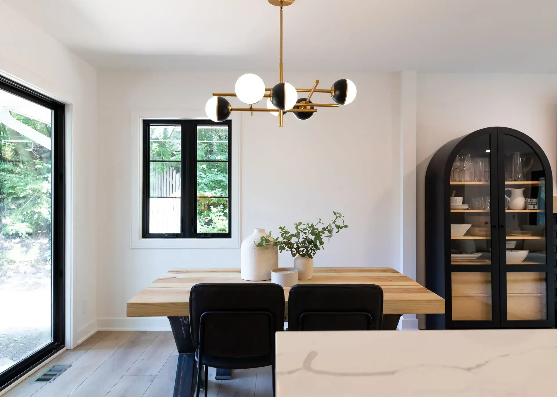 Dining room with wooden table, black chairs, and modern gold chandelier. White walls, large windows.