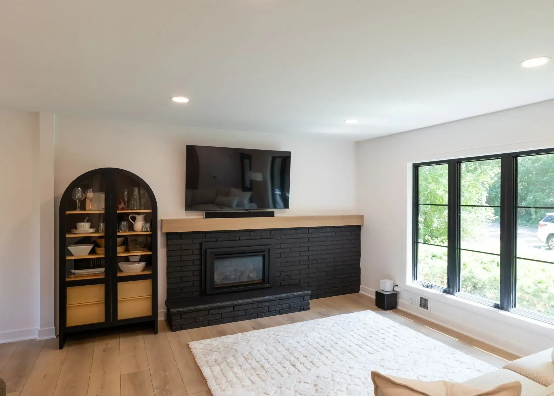 Living room with black fireplace, TV, and black window frames. White walls, wood floor, and rug.