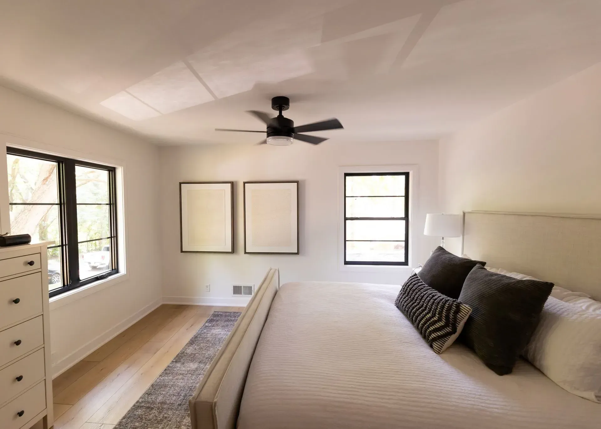 Bedroom with white walls, black windows, and a bed with pillows. Two framed artworks hang above the bed.
