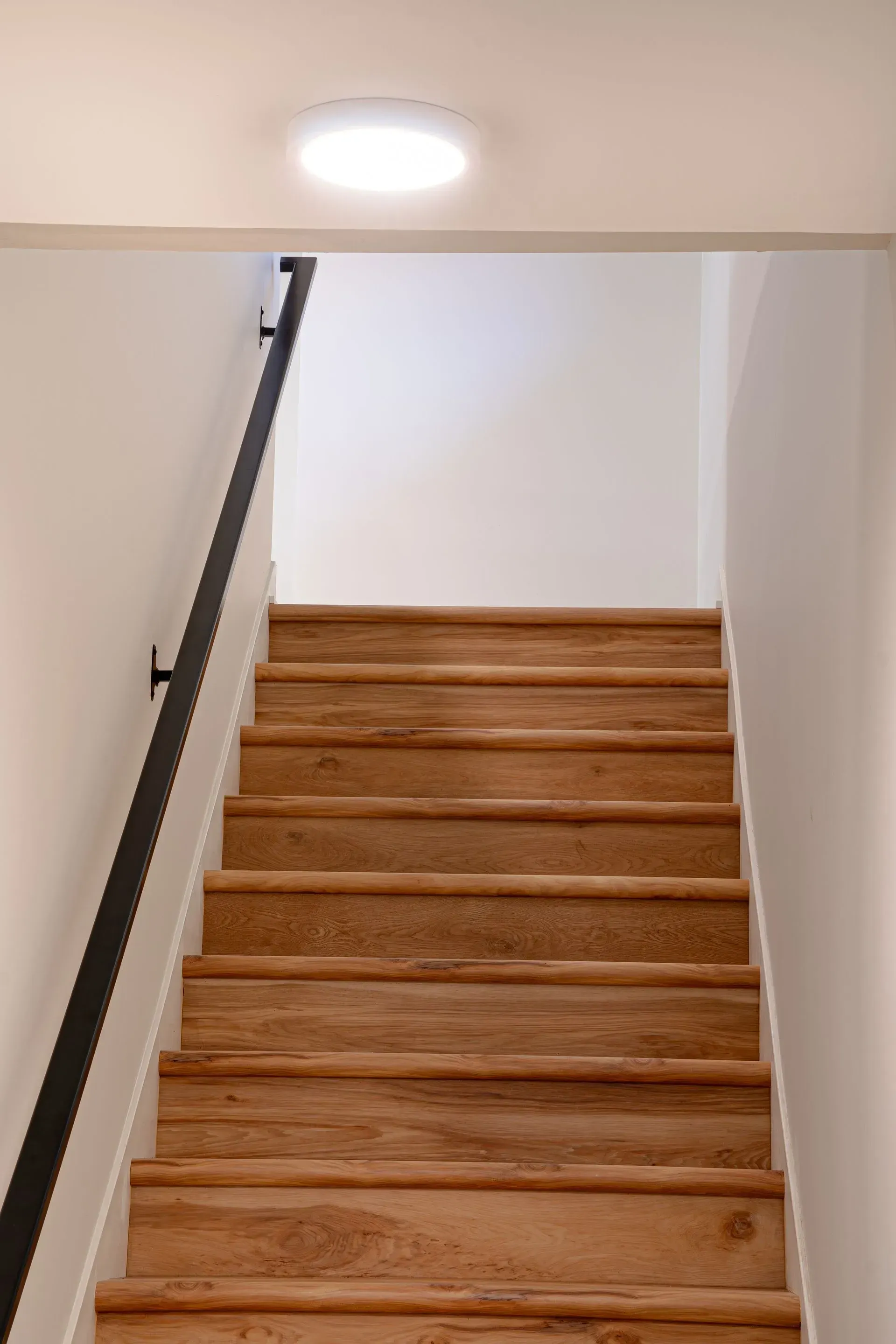 Wooden staircase with black handrail, leading upwards towards a bright, circular ceiling light.