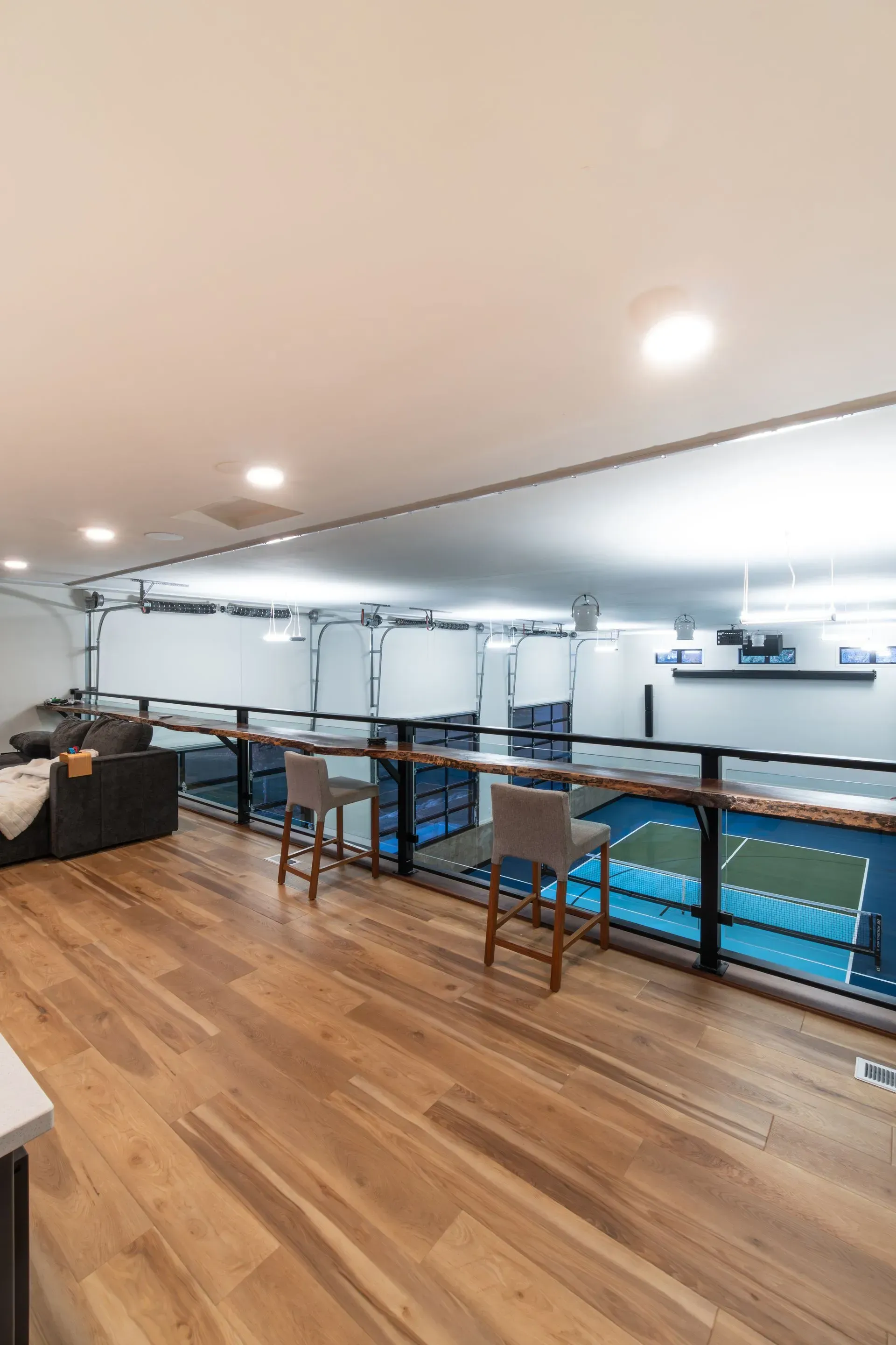 Wooden balcony overlooking an indoor sports court; two bar stools, glass railing.