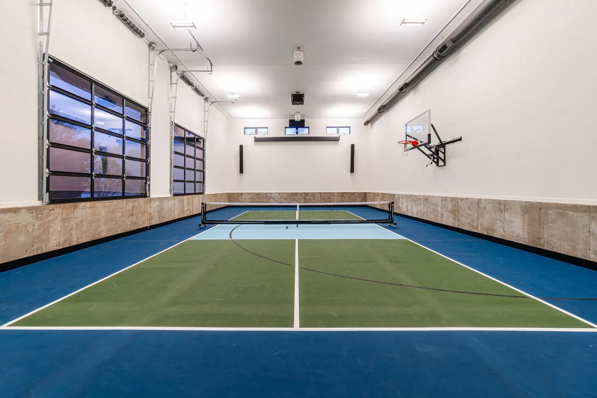 Indoor pickleball court with blue, green, and white surface, net, basketball hoop, and garage door-style window.