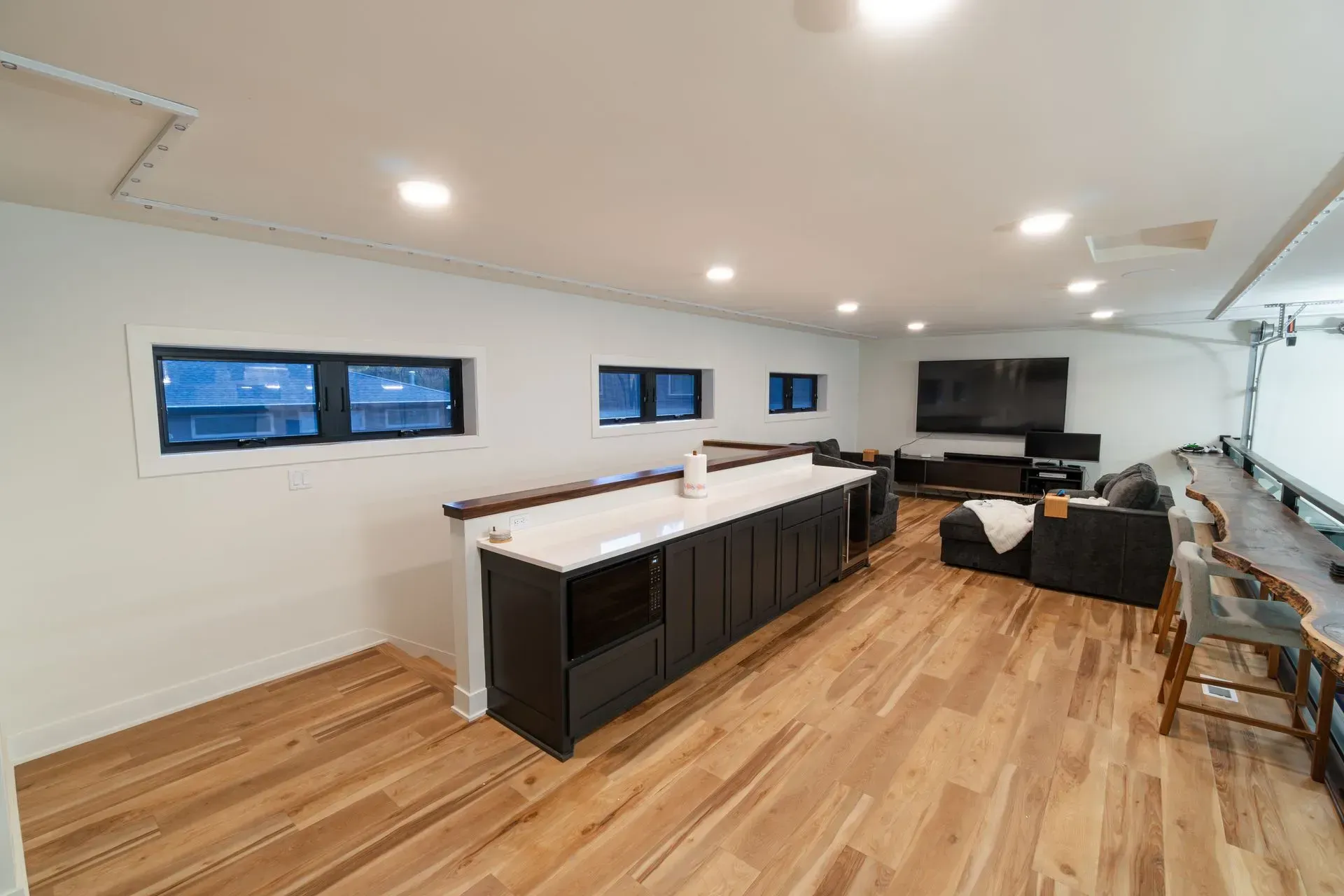 Modern finished basement with bar, TV, and seating. Wooden floors, white walls, and dark cabinets.