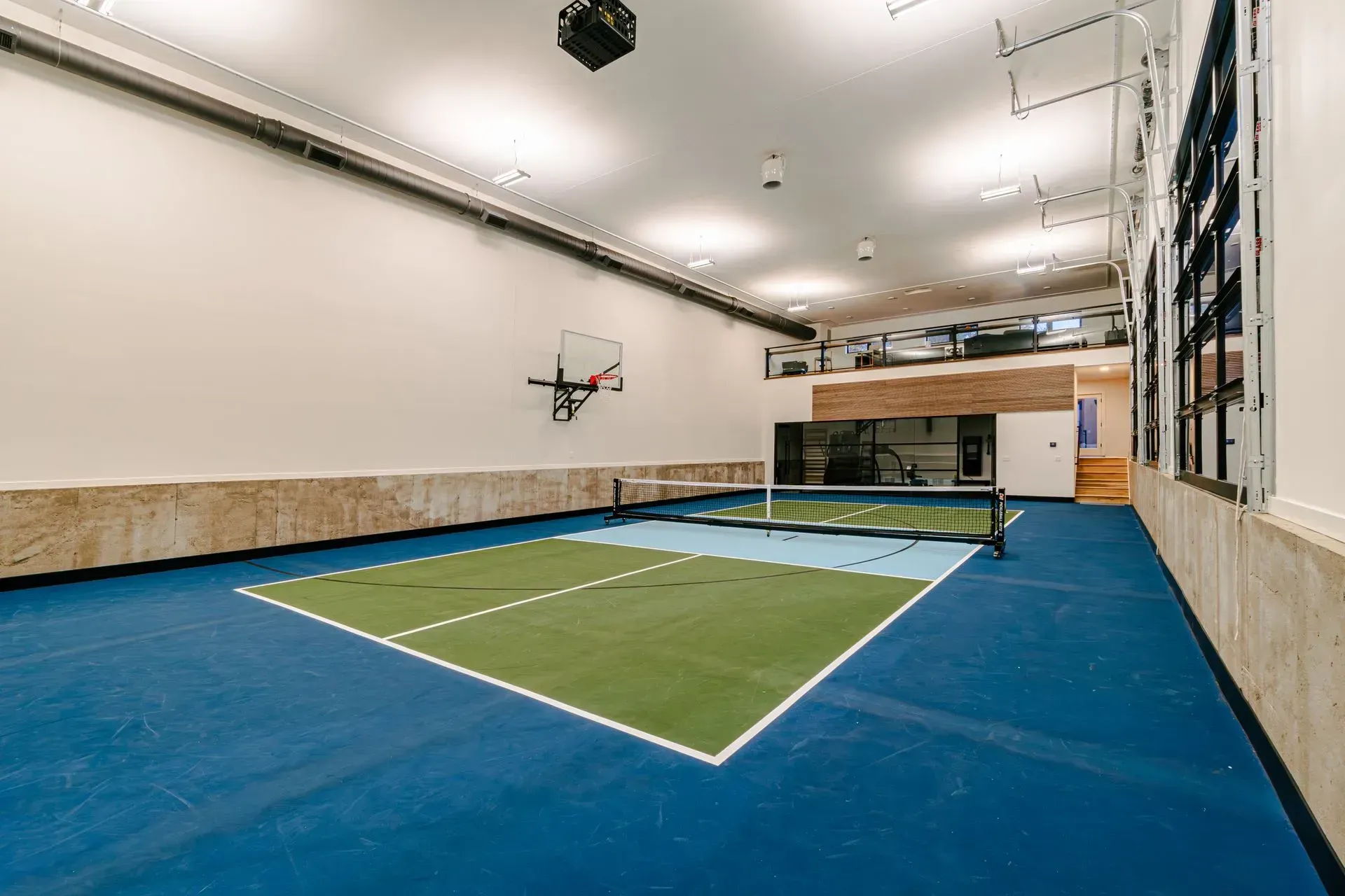 Indoor pickleball court with blue and green flooring, net, basketball hoop, and projector.