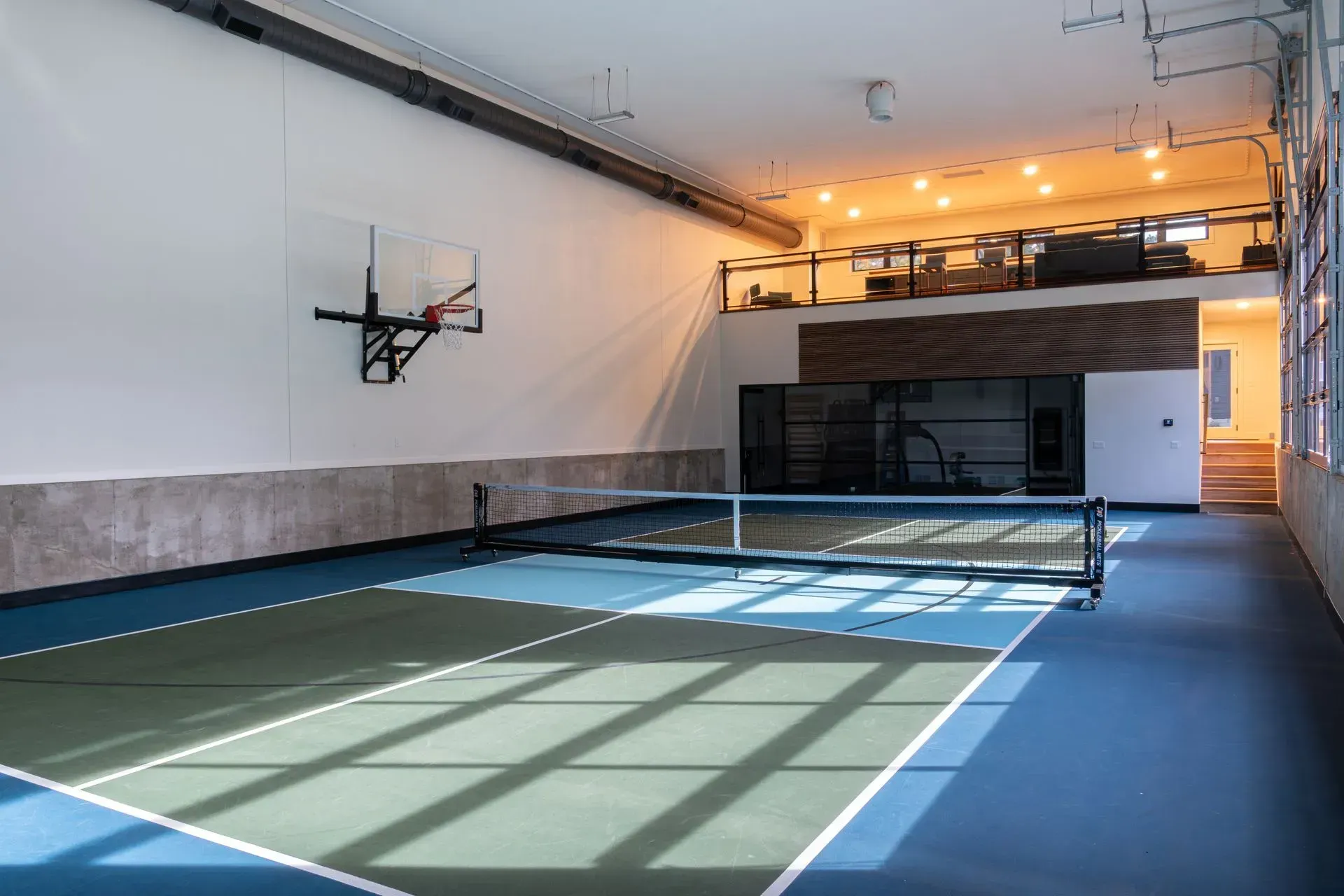 Indoor recreational space with basketball hoop and pickleball court. Blue, green, and gray court with loft area.