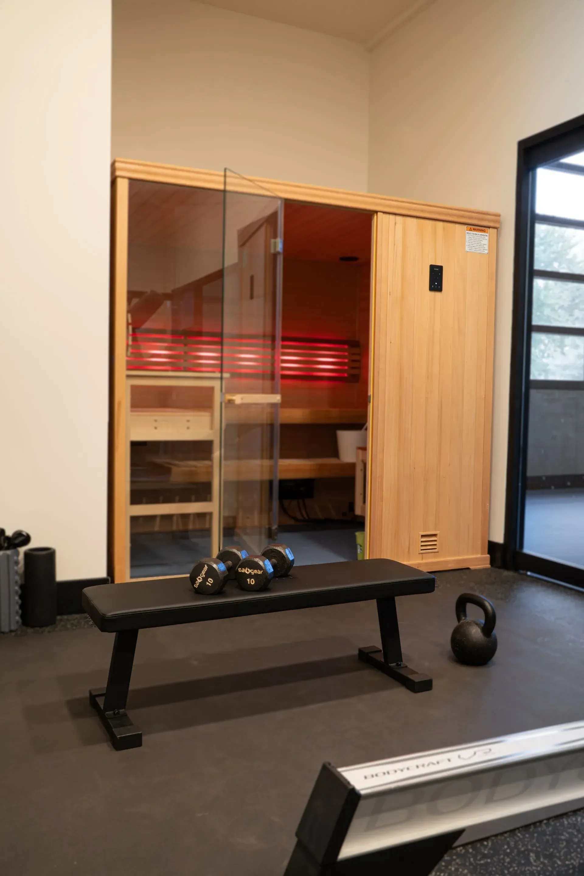A home gym with a sauna, bench, dumbbells, and kettlebell on a rubber floor.