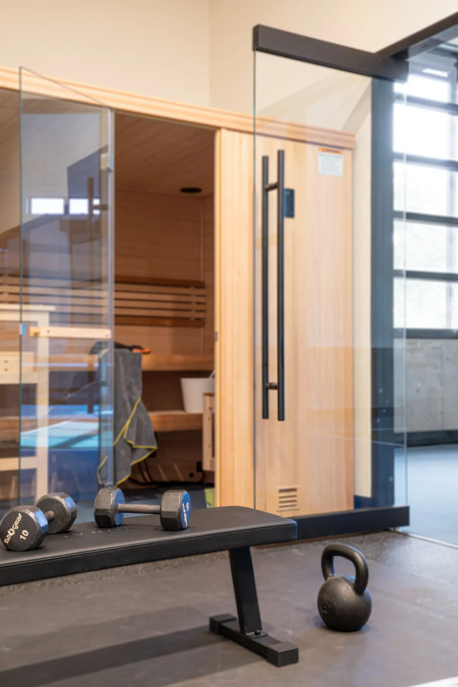 Gym interior with sauna, weights, and bench. Glass doors and light wood sauna.