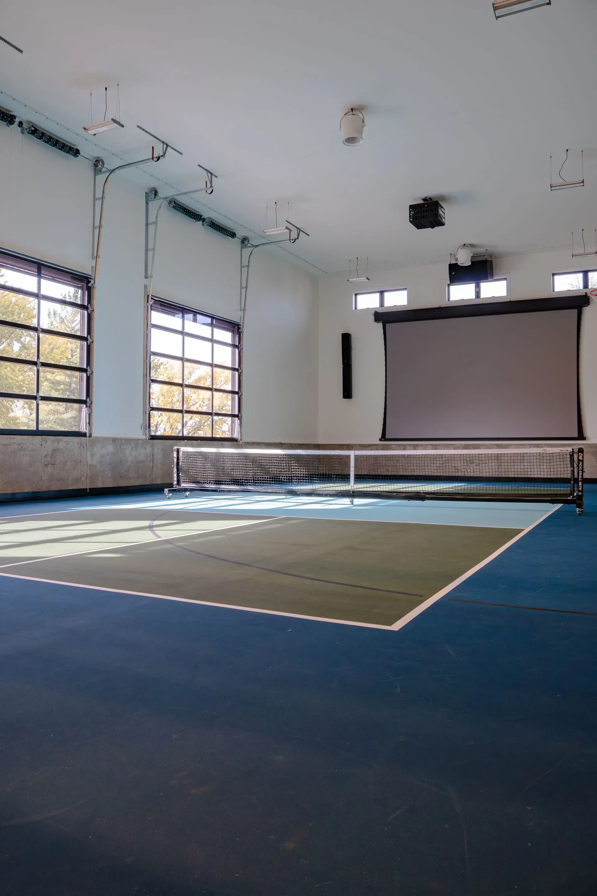 A blue and green pickleball court inside a bright room, net set up, screen hanging, windows on the left.