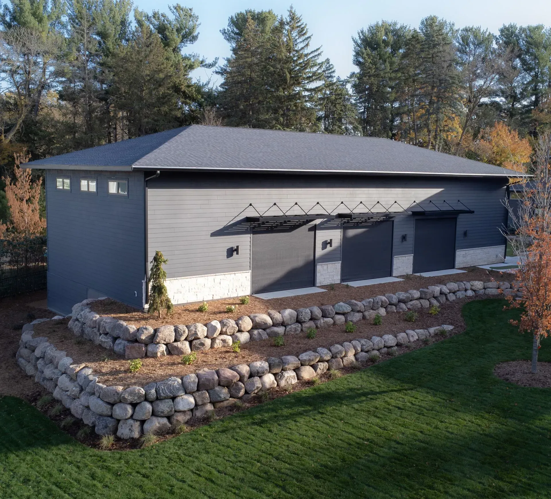 Gray building with garage doors, rock retaining wall, and a green lawn.