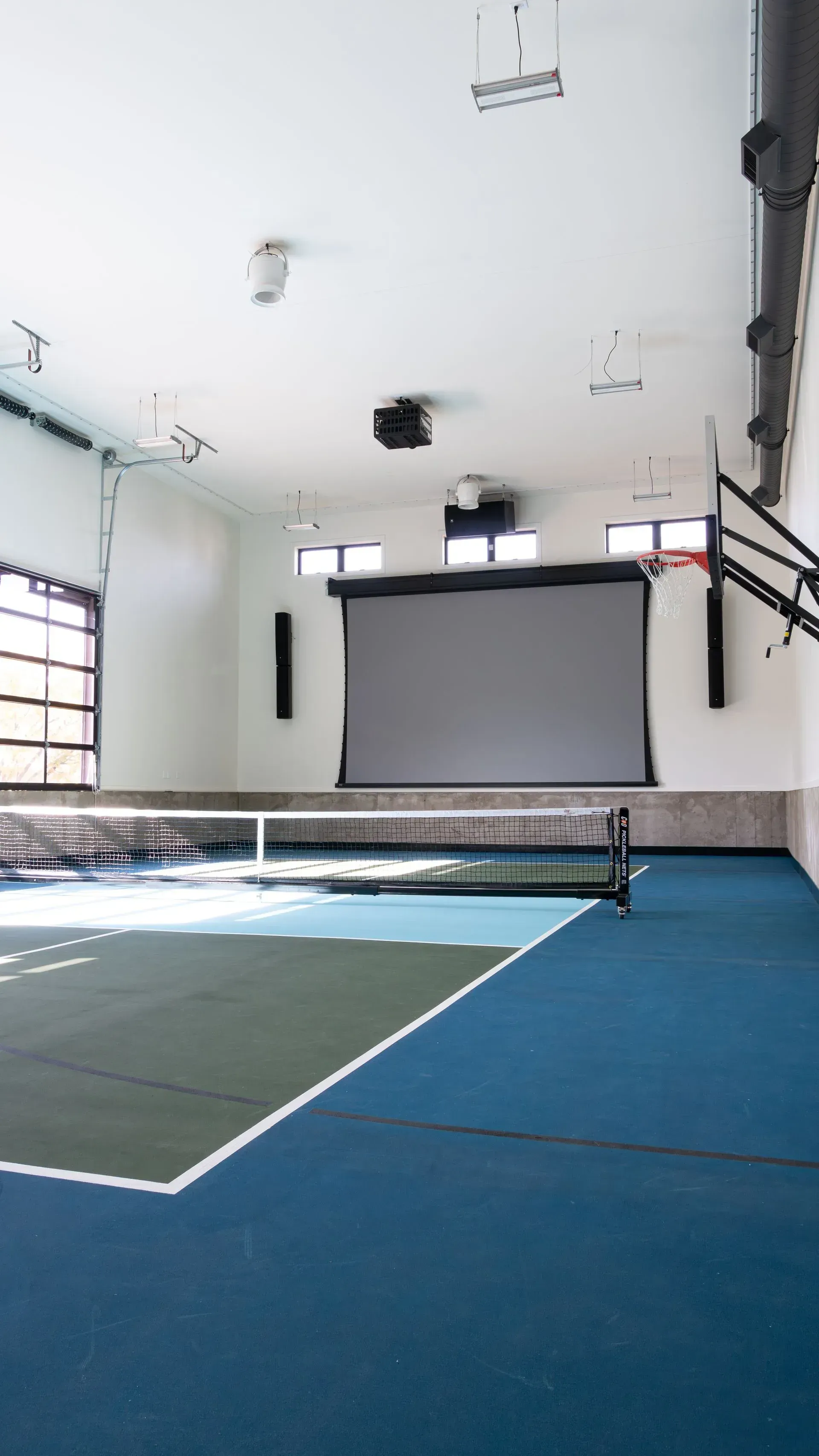 Indoor sports court with blue and green flooring. Projection screen, speakers, basketball hoop, and garage door visible.