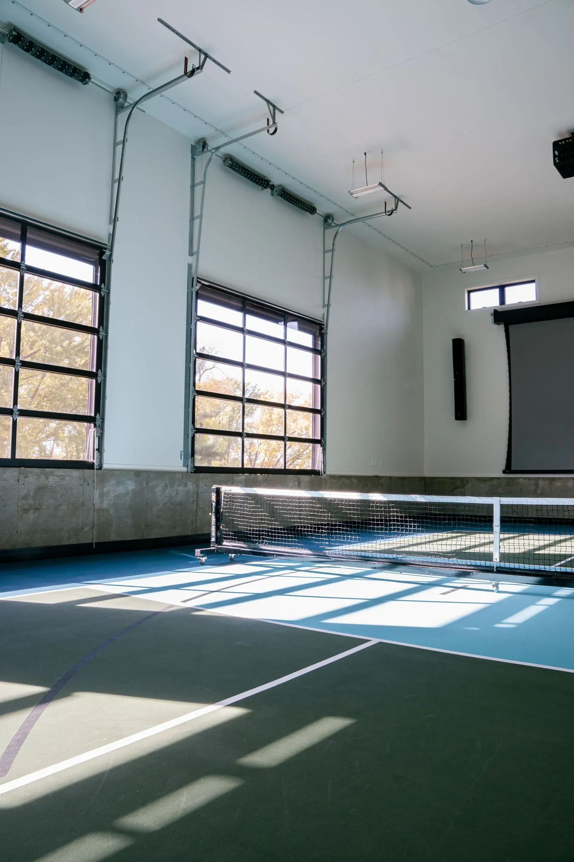 Indoor pickleball court with net, dark green and blue court, large windows, and projection screen.