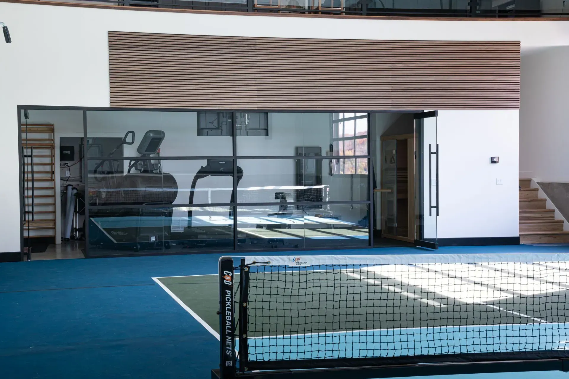 Gym with treadmills and equipment viewed through glass wall on blue court, net in foreground.