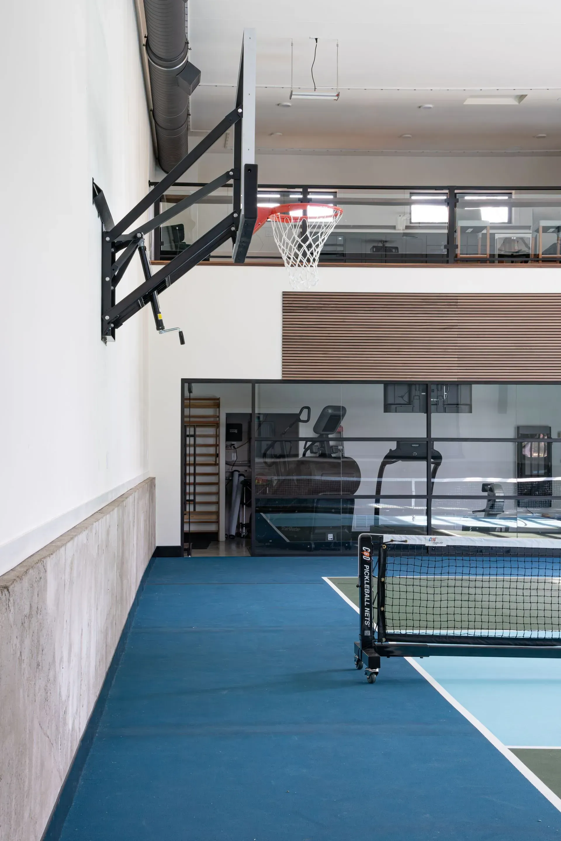 Indoor basketball court with hoop on a white wall, blue floor, and equipment visible through glass.