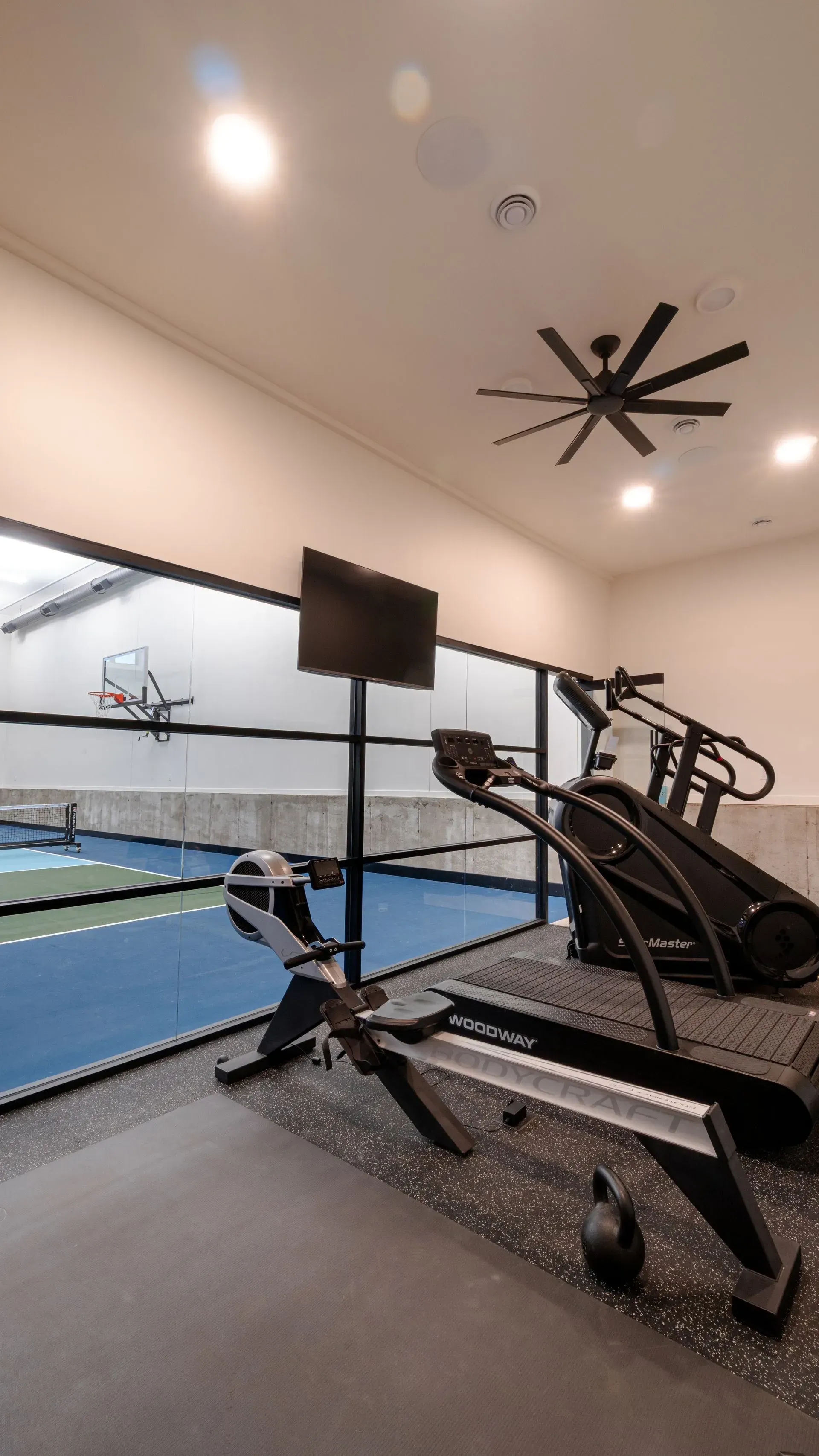 Gym with treadmill, rowing machine, TV, and view of a sports court through a window.