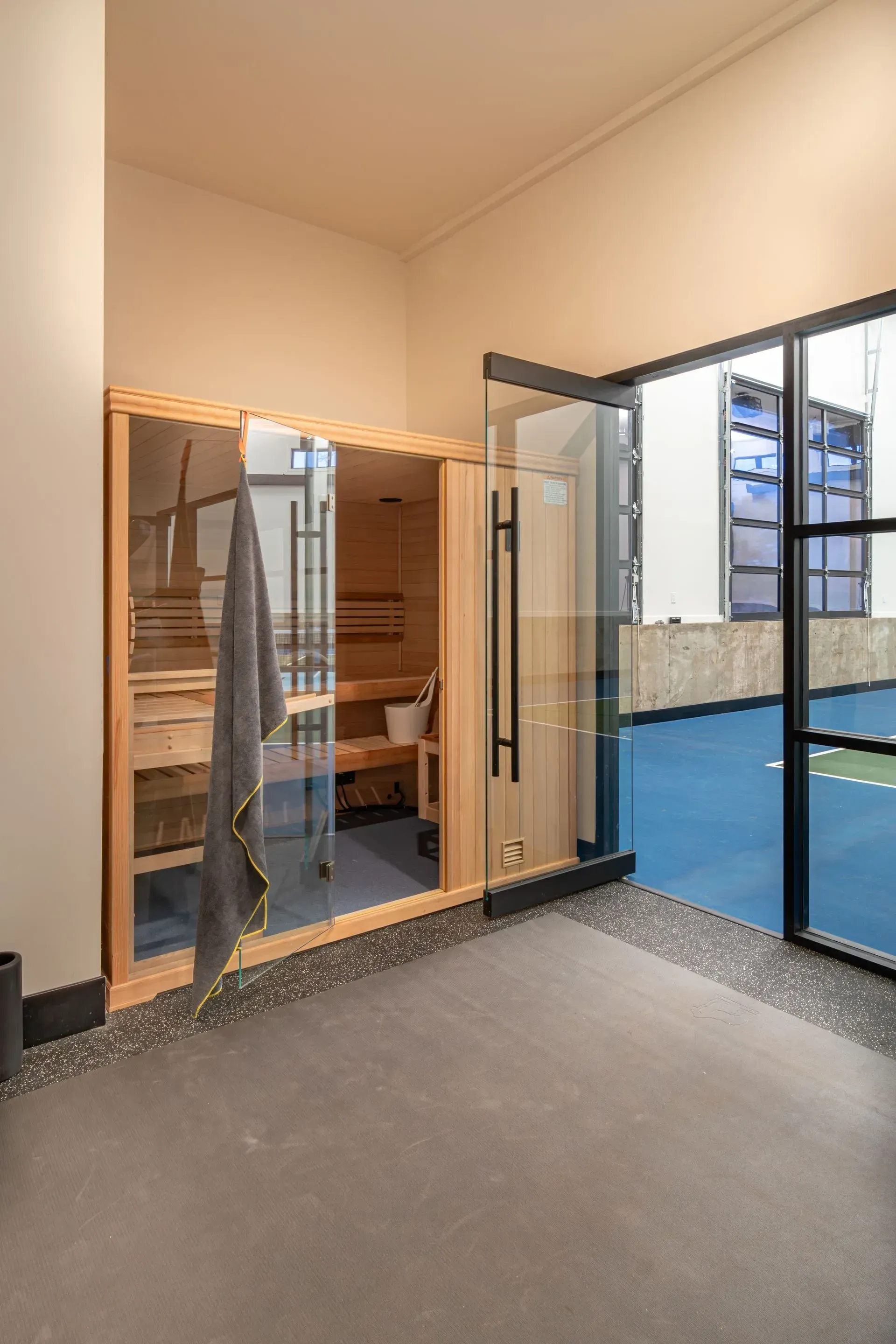 Sauna with glass door, adjacent to an open door overlooking a blue court. Gray towel hangs on the sauna door.