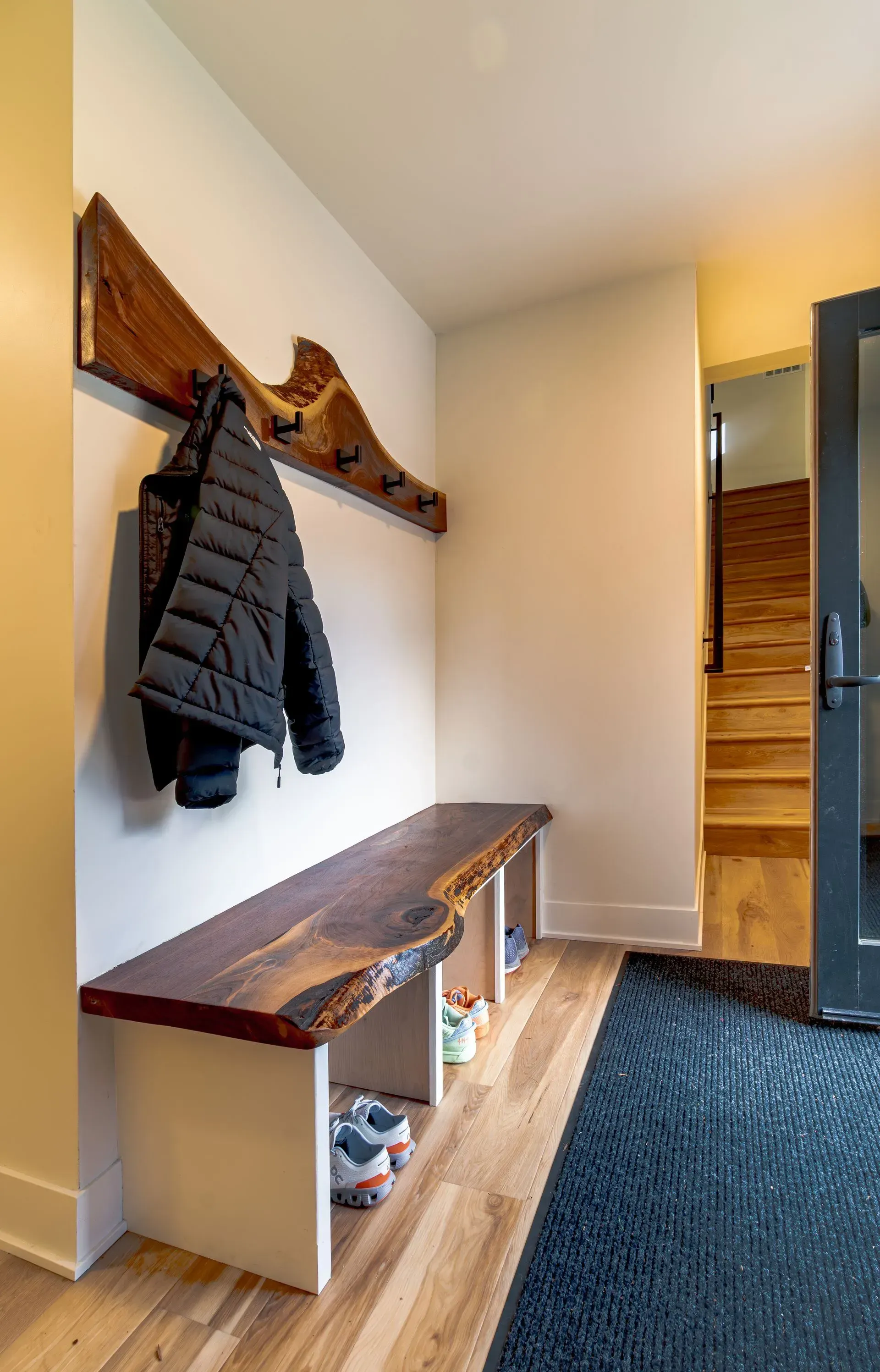 Entryway with bench, coat rack, shoes, and stairs. Dark wood accents contrast with light walls.