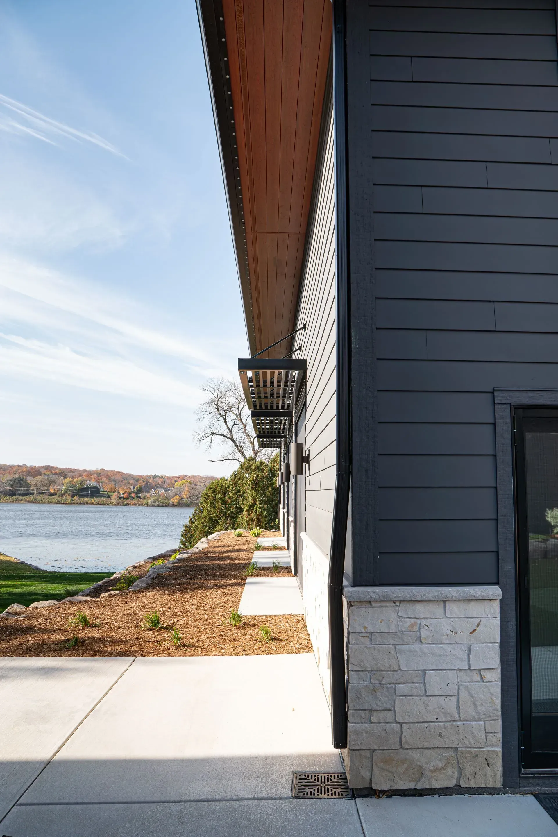 Exterior view of a modern gray house, with a stone foundation, overlooking a river on a sunny day.