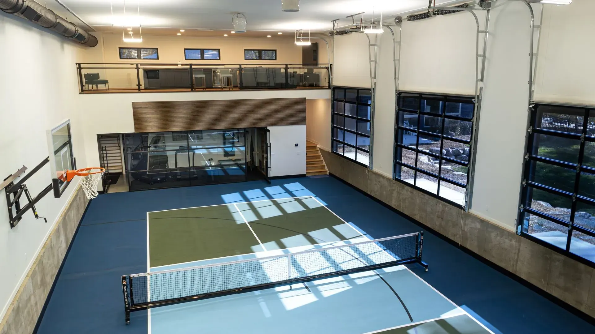 Indoor pickleball court with blue and green playing surface, glass garage doors, and a balcony.