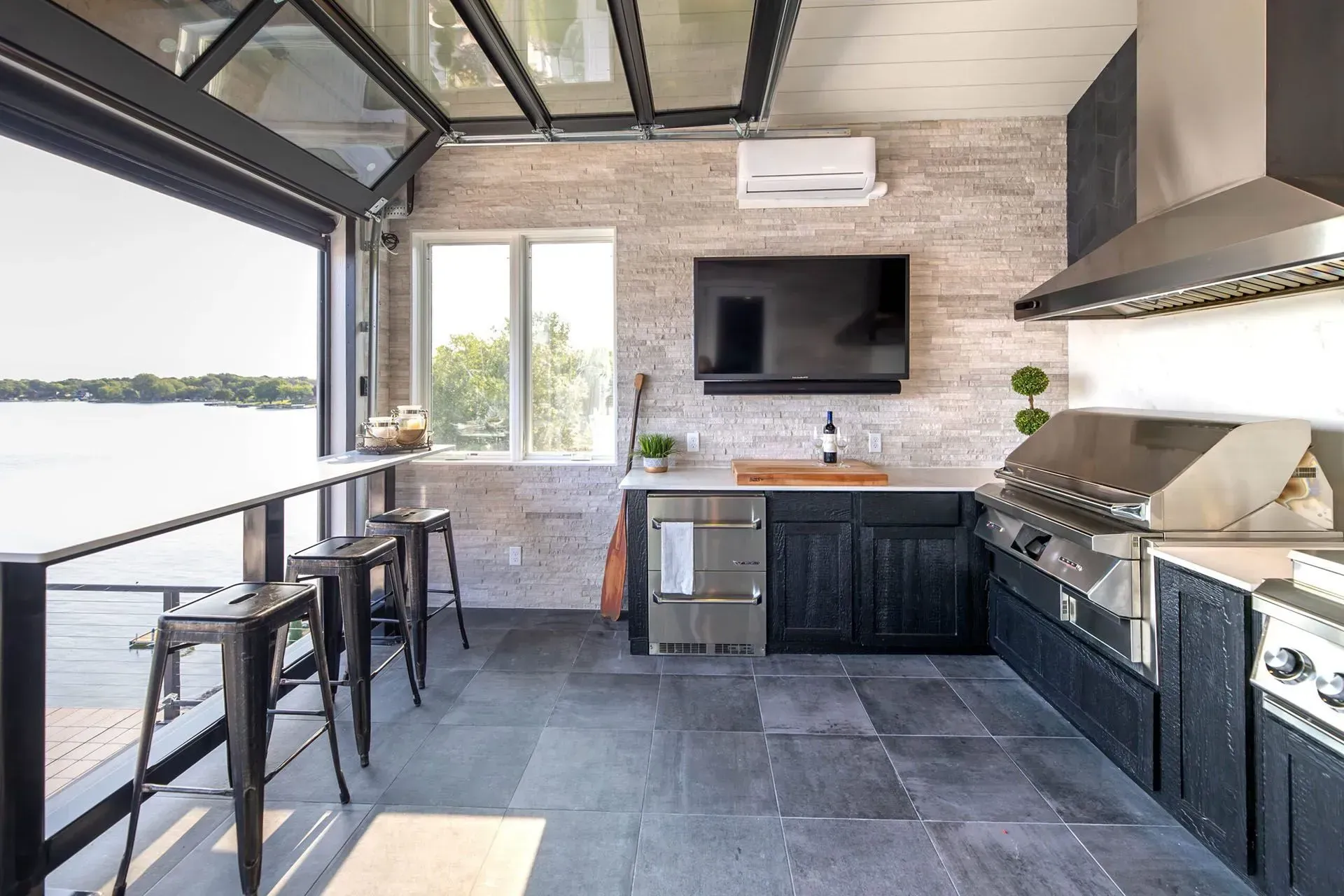 Outdoor kitchen with bar seating, grill, TV, and open doors overlooking water.
