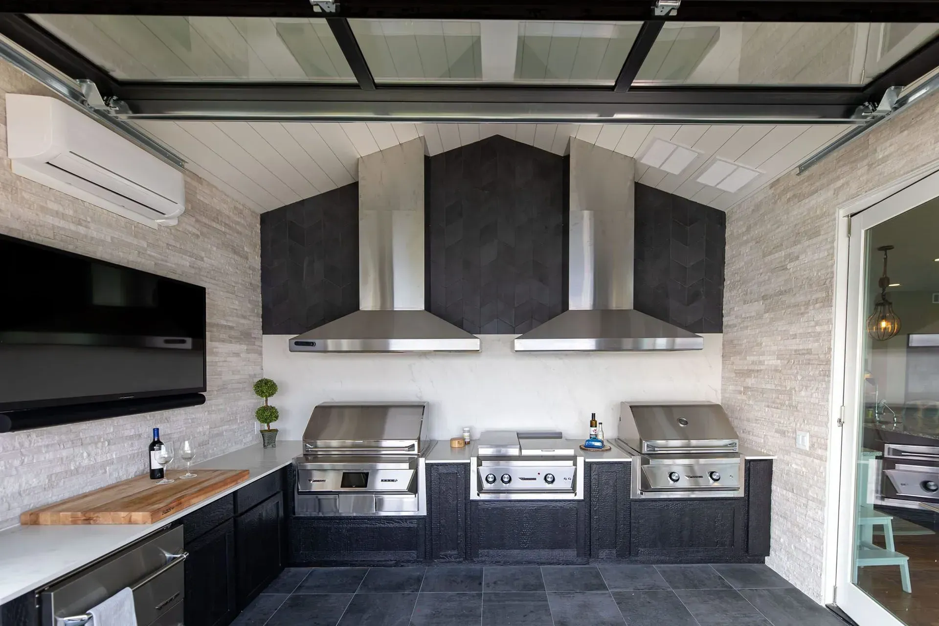 Outdoor kitchen with stainless steel grills, range hoods, and a wall-mounted TV.
