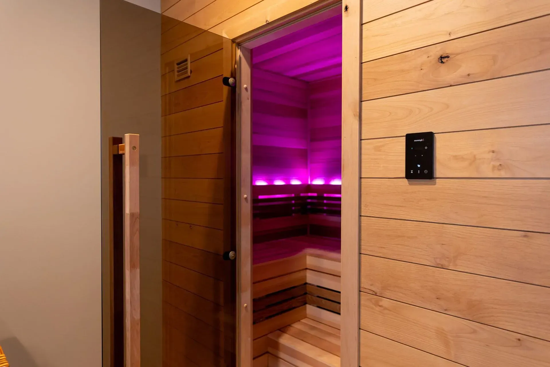 Wooden sauna interior with open door, lit by bright pink lights.