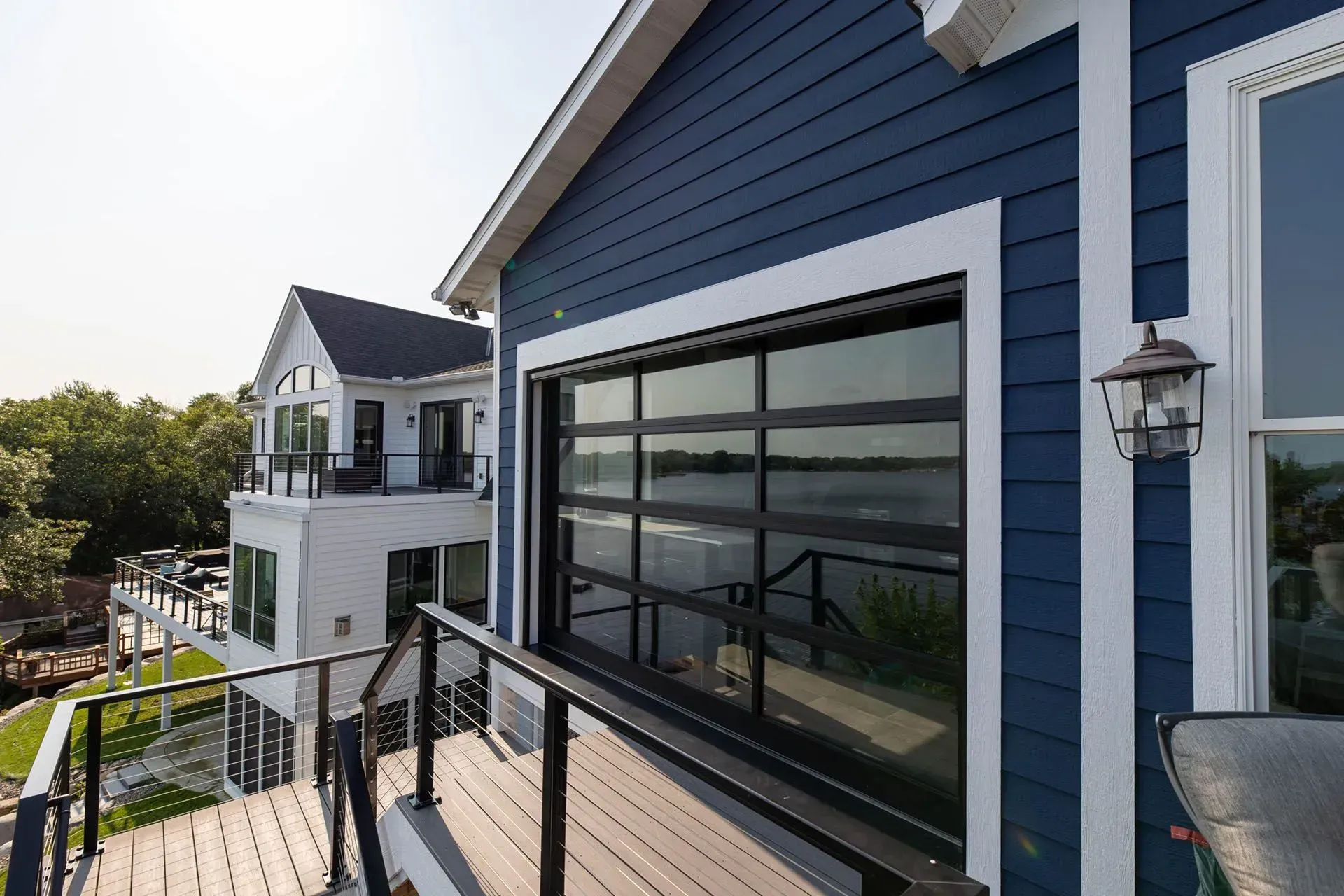 Blue building with glass garage door facing water, deck in foreground, another house in the background.