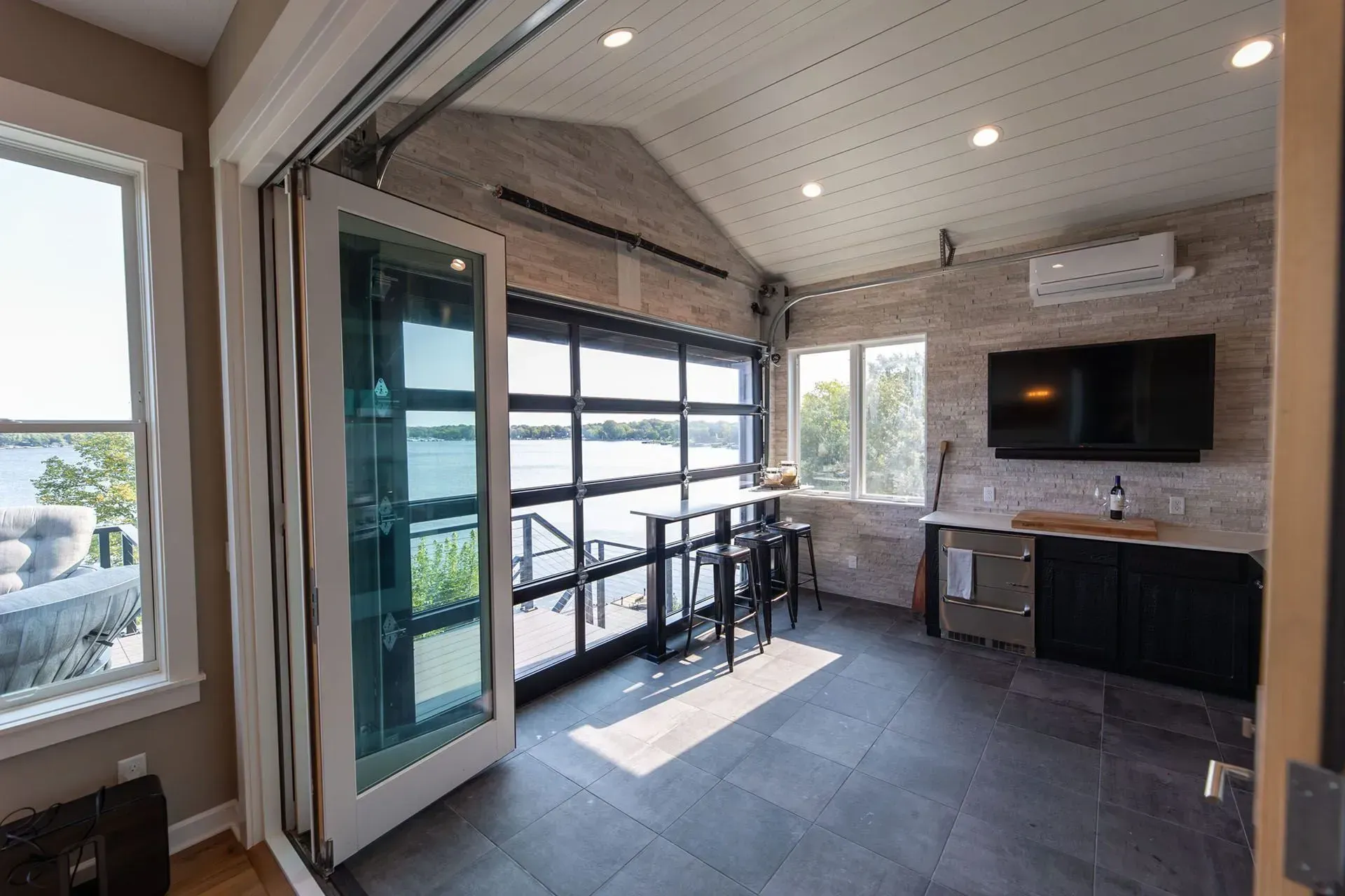 Interior of a sunroom with large windows facing a lake. Features bar, TV, and garage door.