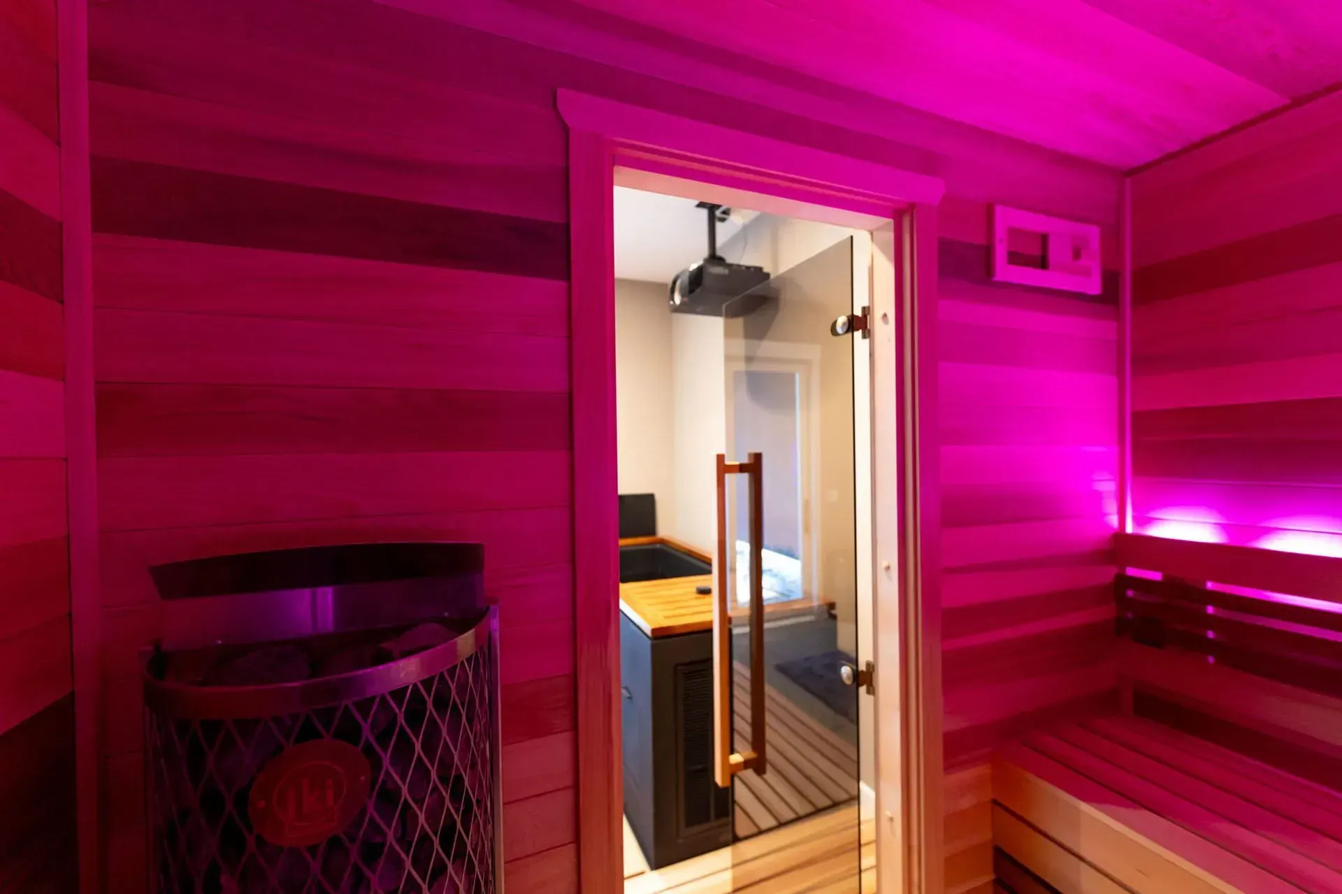 Interior of a sauna with pink lighting, wooden benches, and an open doorway to another room.