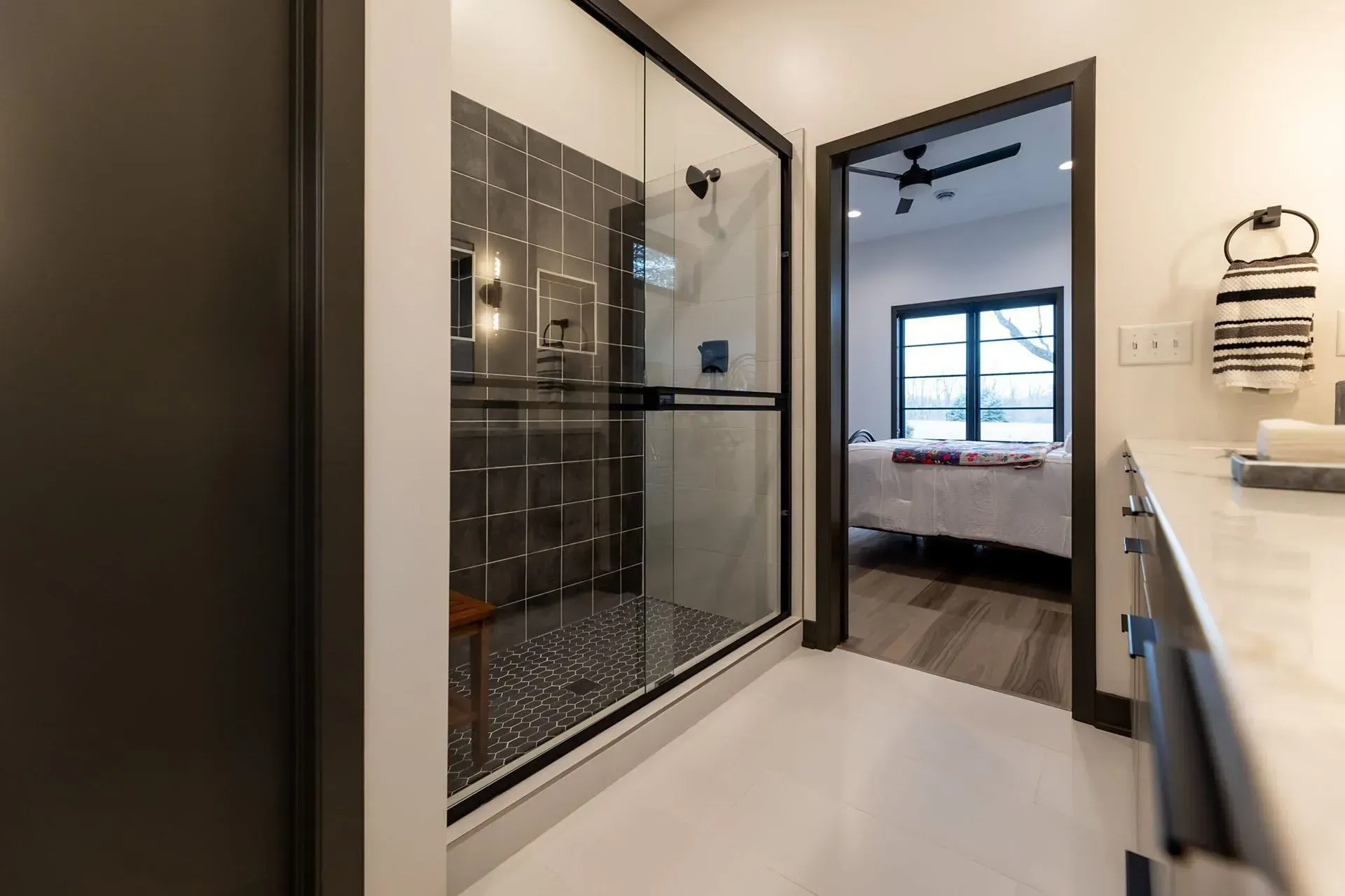 Modern bathroom with a glass-enclosed shower, dark tiles, and doorway leading to a bedroom.