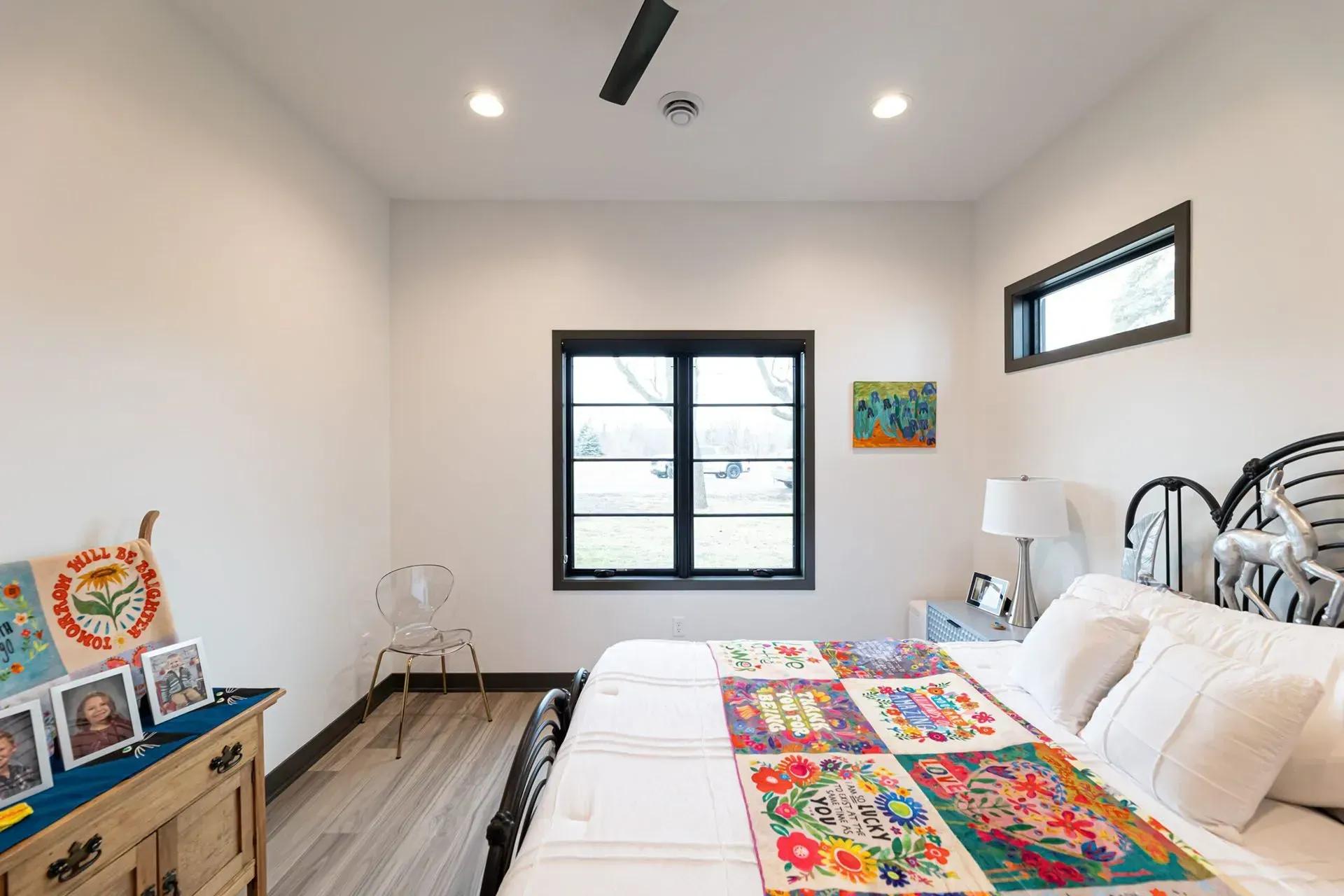 Bedroom with white walls, black-framed windows, bed with colorful quilt, dresser, and small art.