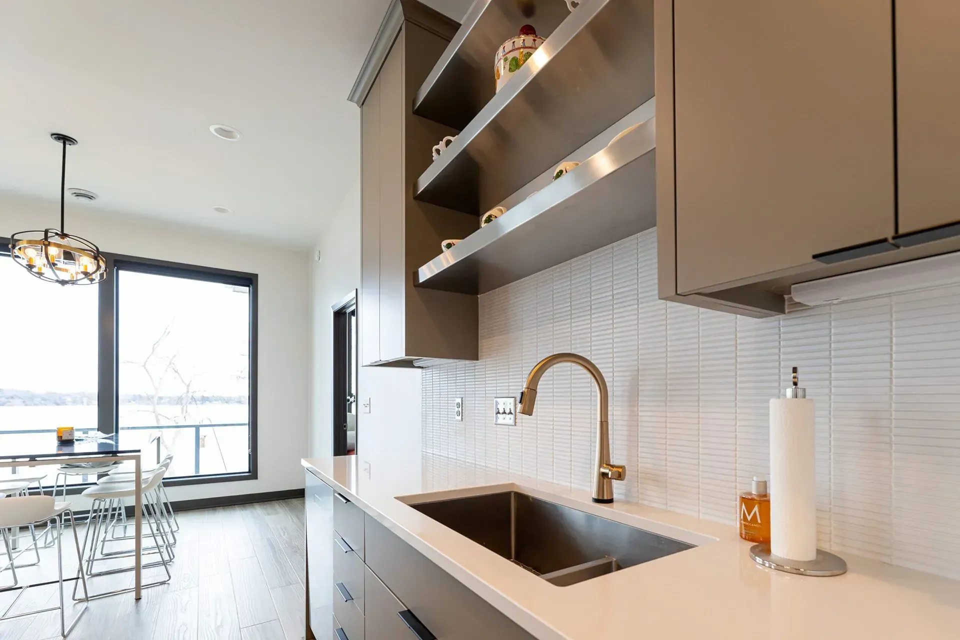 Modern kitchen with stainless steel sink, gold faucet, open shelving, and large window overlooking a waterscape.