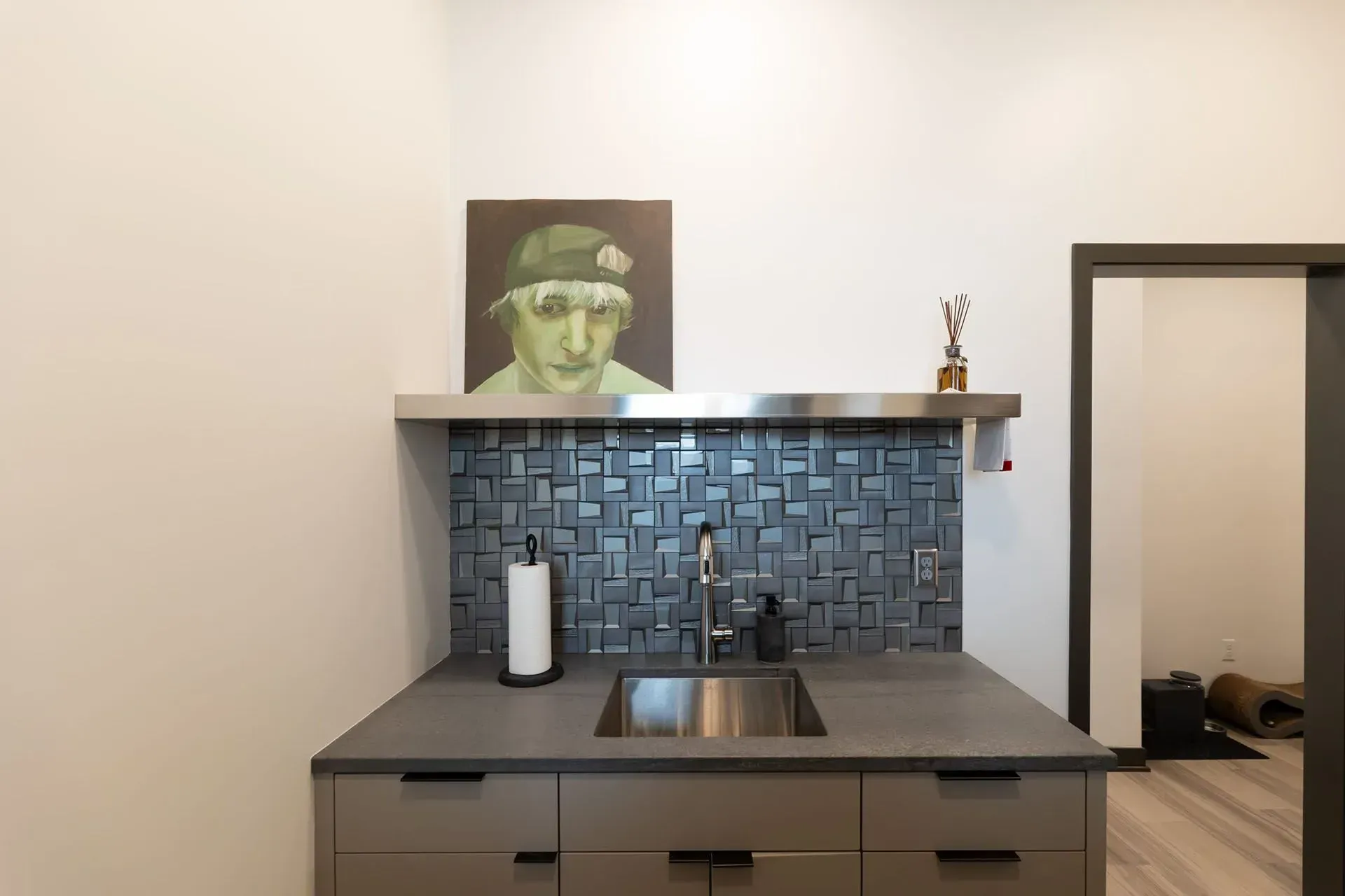 A wet bar with a dark gray countertop, sink, and backsplash, with a painting above and an open doorway.
