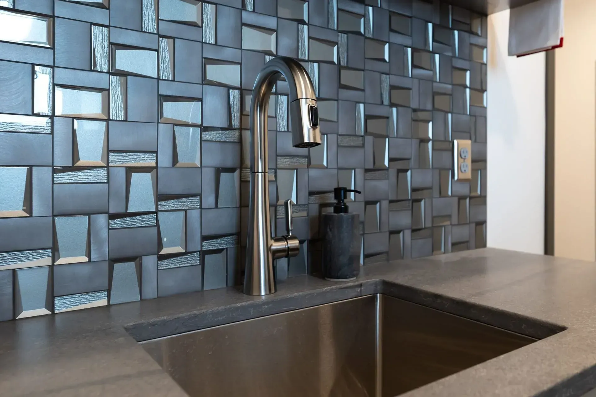 Kitchen sink with stainless steel faucet, dark soap dispenser, and textured blue tile backsplash.