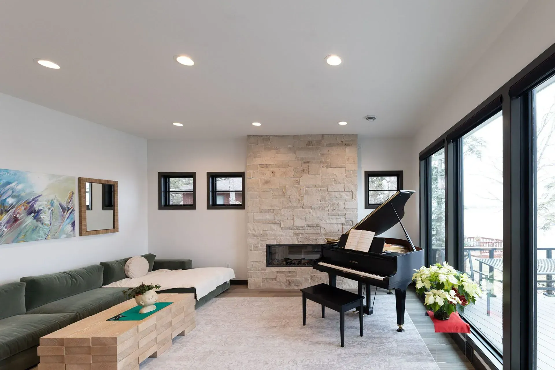 Living room with grand piano, fireplace, large windows overlooking a deck, green sofa, and light wood coffee table.