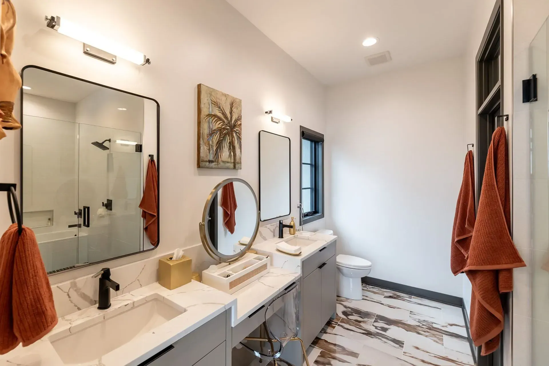 Modern bathroom with white walls, weathered wood floor, and two sinks with black fixtures; rust-colored towels.