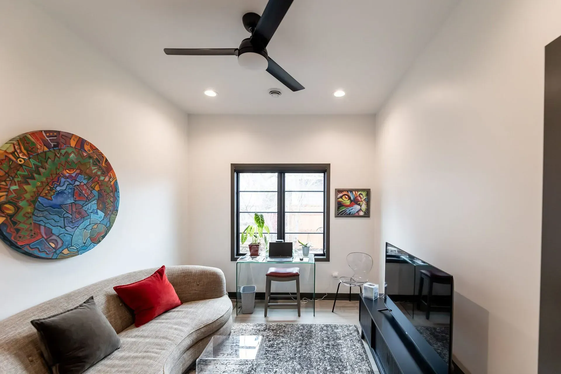 Living room with white walls, sofa, desk by window, artwork, black TV, ceiling fan.