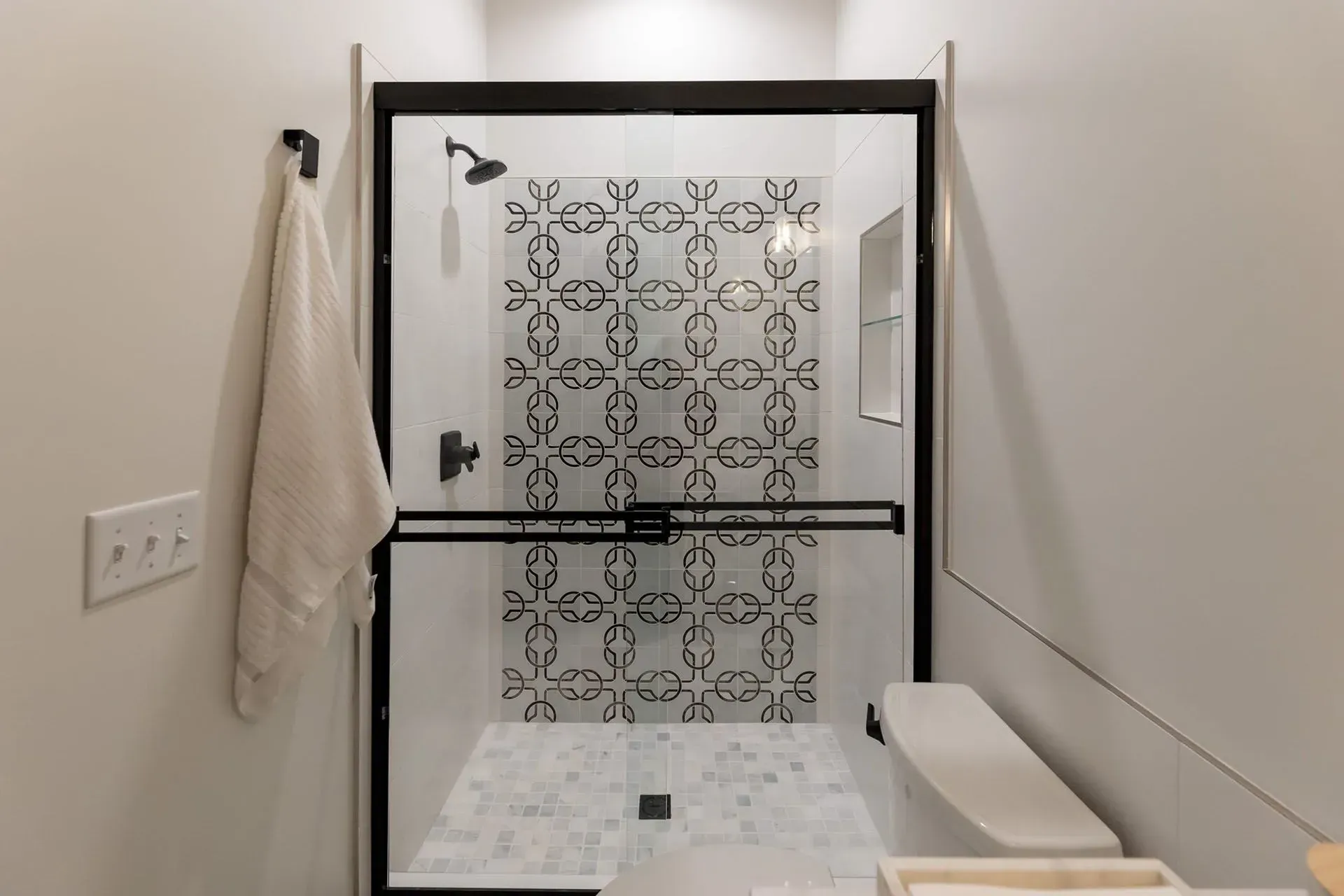Modern shower with black-framed glass door, white tiles, patterned accent wall, and a towel hanging on the left.
