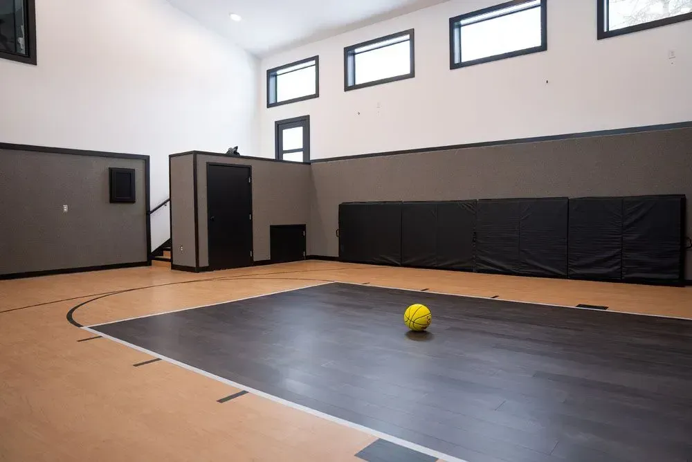 Indoor basketball court with yellow ball on dark floor, padded wall, small windows.