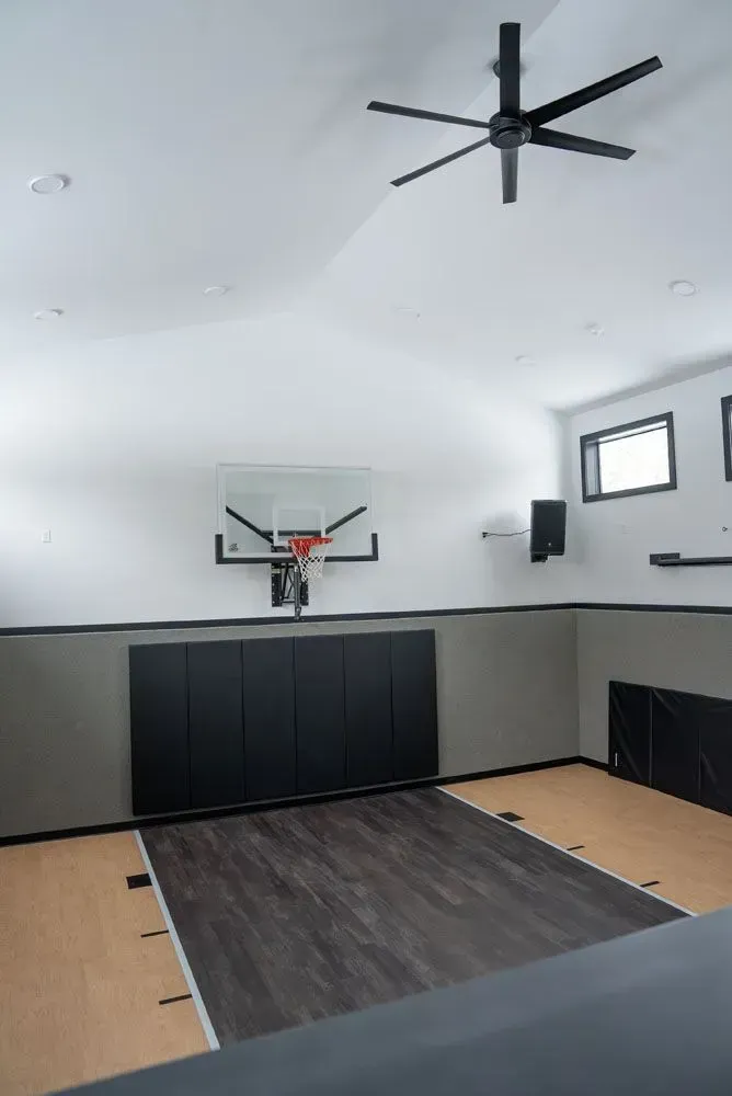 Indoor basketball court with hoop, black padding, and ceiling fan. Light wood and gray walls.